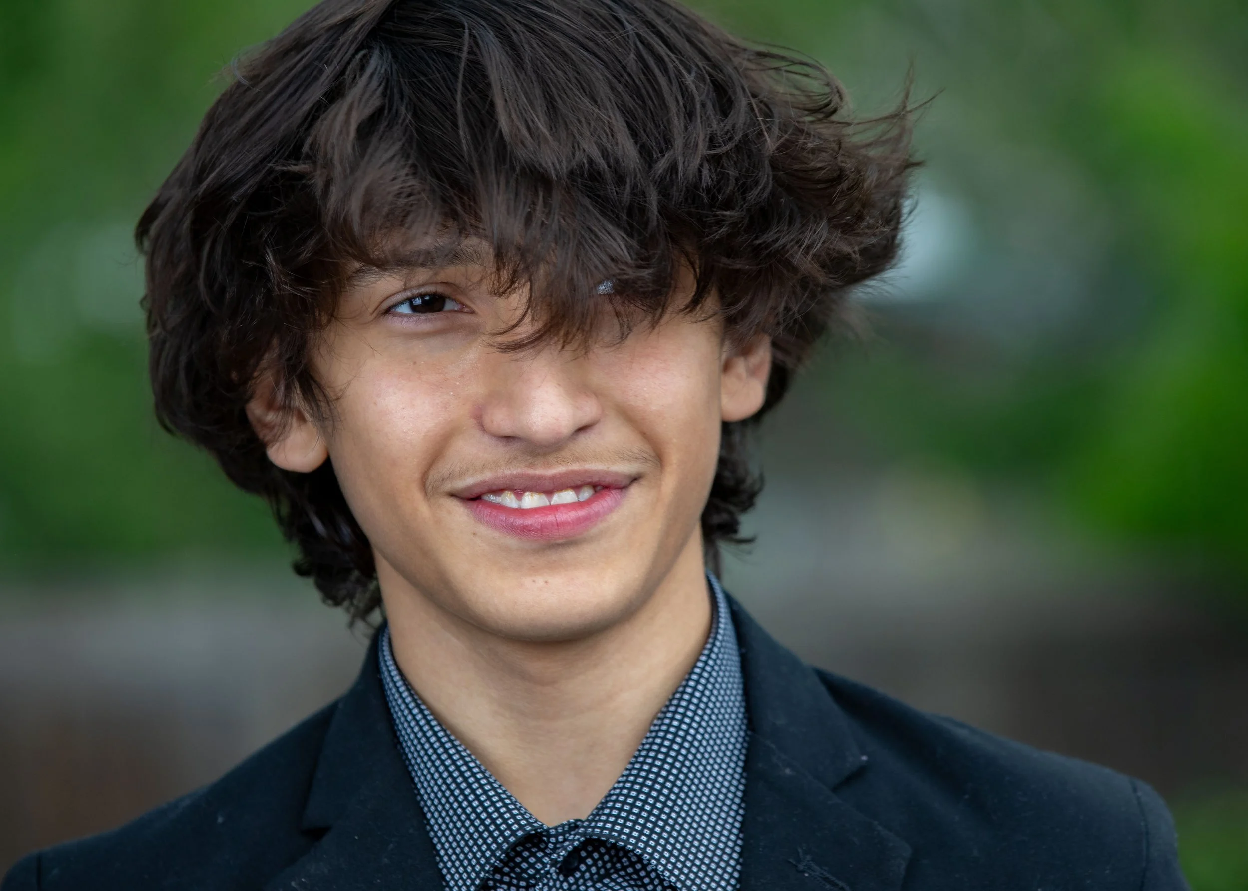 A  photo of a young man with curly brown hair, wearing a black blazer and a patterned shirt, smiling outdoors.  Family child children portrait in park trees teenager photography Family portraits in Denver