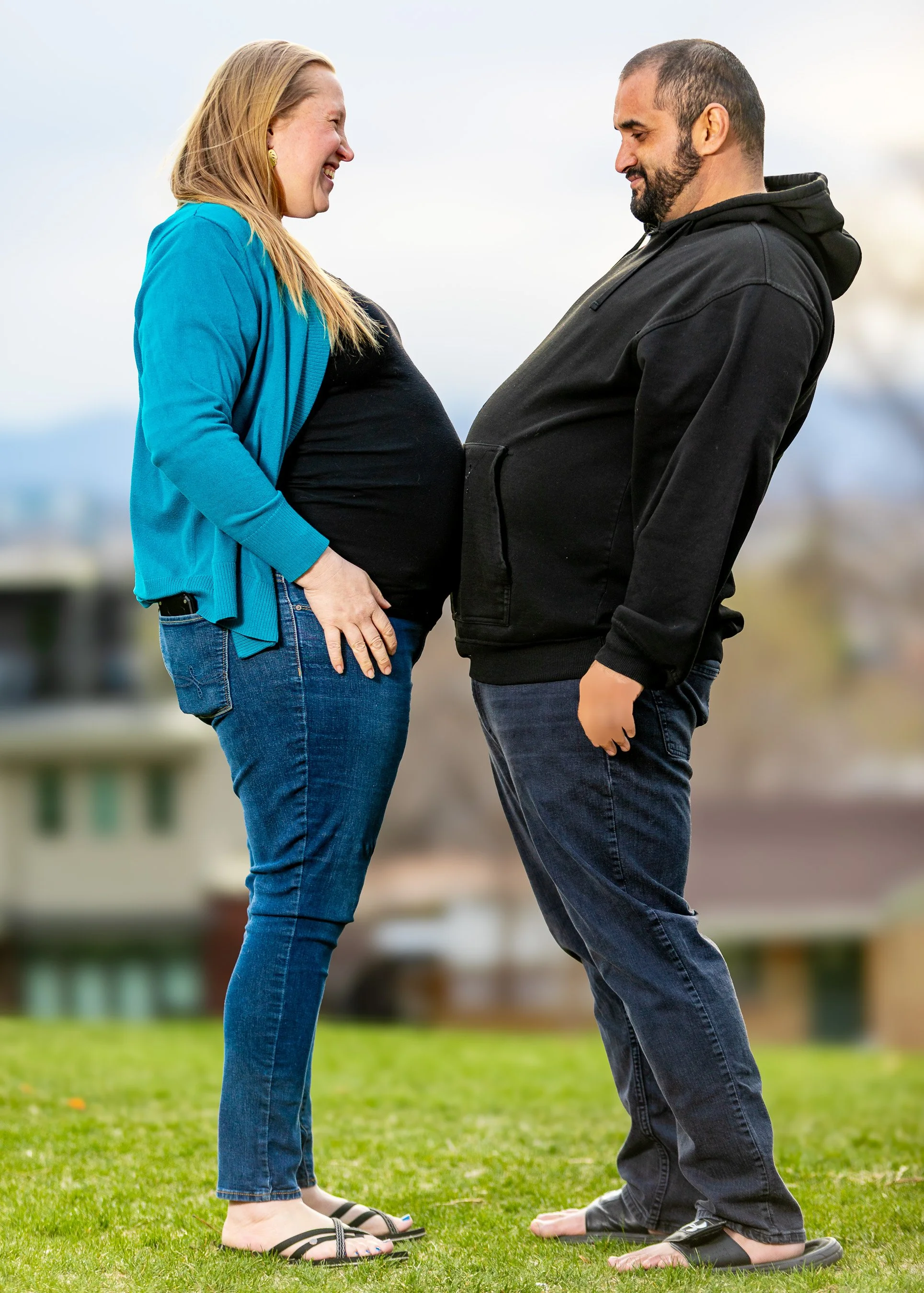 A pregnant woman smiling and looking at a man, both kneeling on grass outdoors, holding hands, facing each other, with a blurred background of trees and buildings.  Pregnancy and maternity couple photography in Denver Colorado