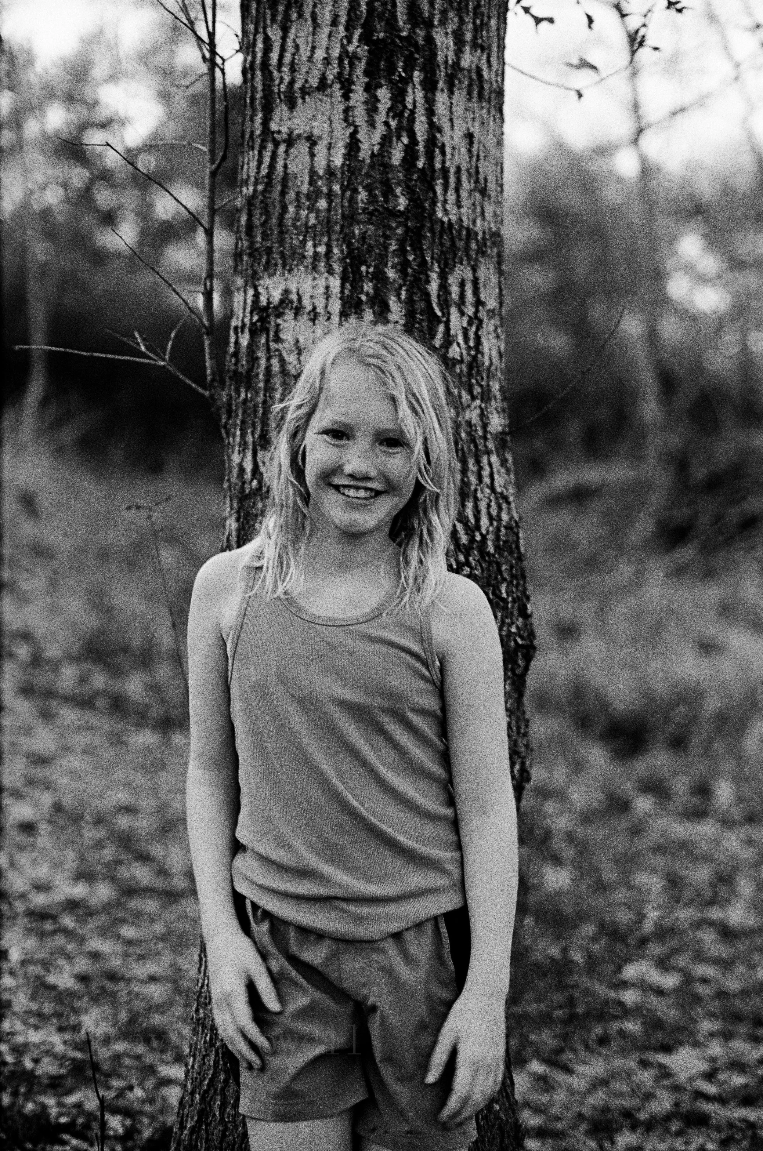 Black-and-white film portrait of a smiling girl standing beside a tree outdoors.