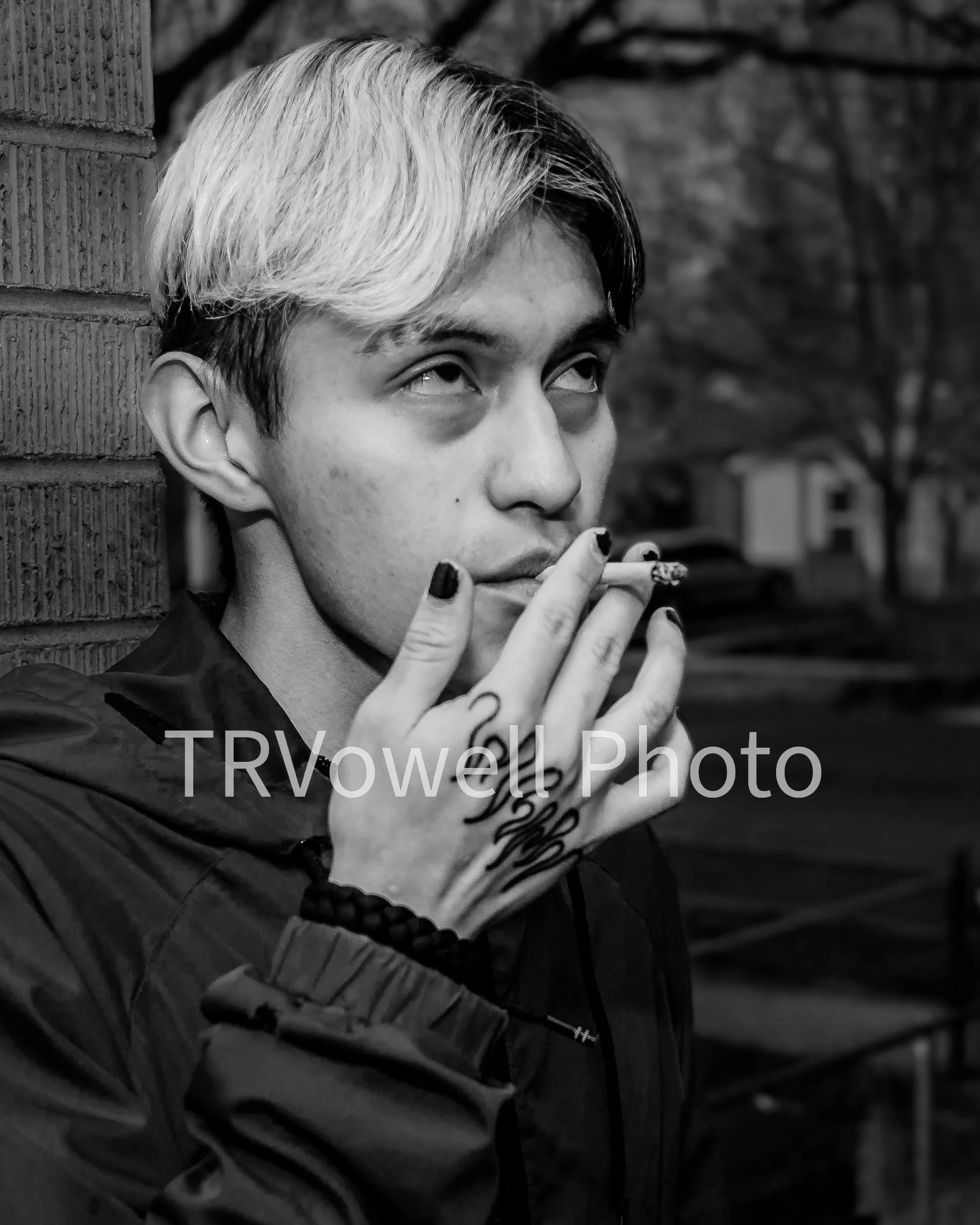 Black and white photo of a young man with two-toned hair, smoking a cigarette, leaning against a brick wall, with tattoos on his hand, wearing a dark jacket and a beaded bracelet.