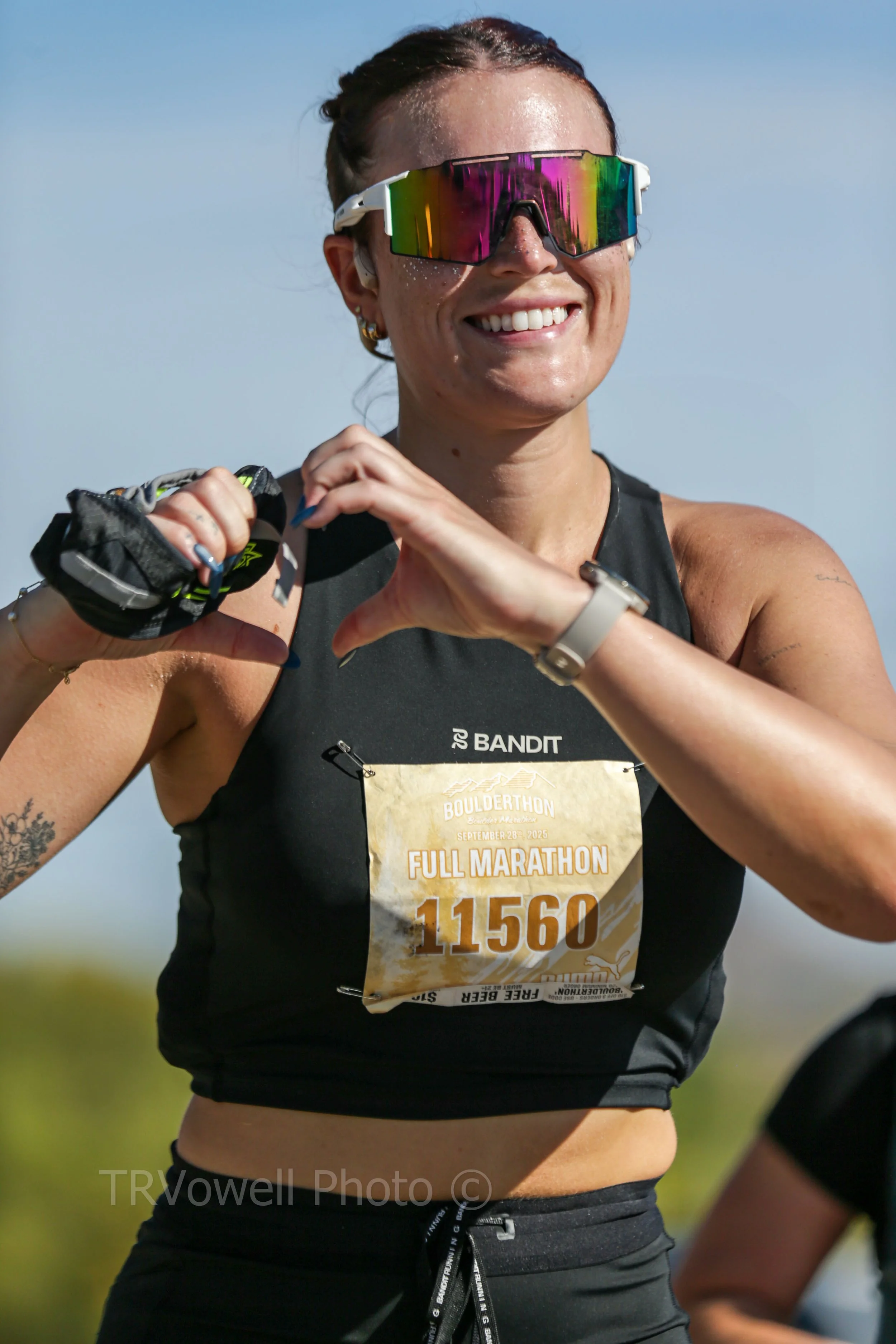 A smiling woman wearing reflective sunglasses, a black sports tank top, and black running shorts, making a heart shape with her hands during a marathon event.