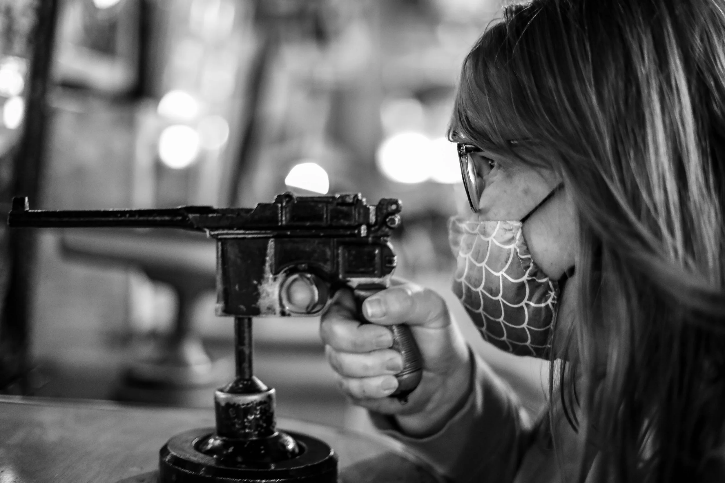 A portrait of woman wearing glasses and a face mask, holding a pistol-shaped object, aiming and looking intently at her target.  Documentary editorial style candid Black and White photography 