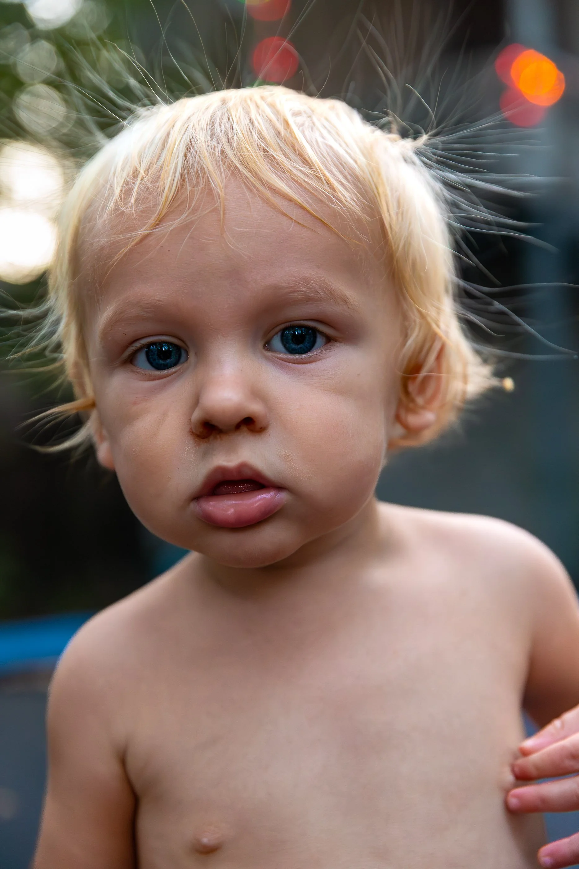 Close-up of a young boy with blue eyes, blonde hair, and fair skin looking directly at the camera. The background is blurred with colorful lights.