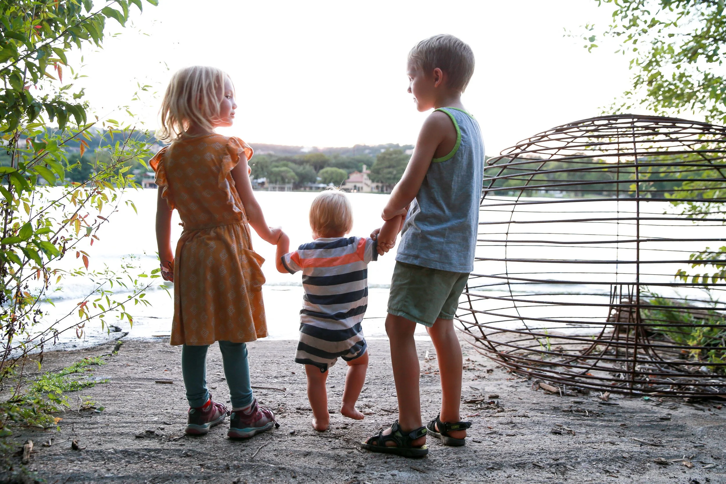 Four children standing on a sandy ground near a body of water, holding hands and facing the water, with trees and a house in the background, during daylight.