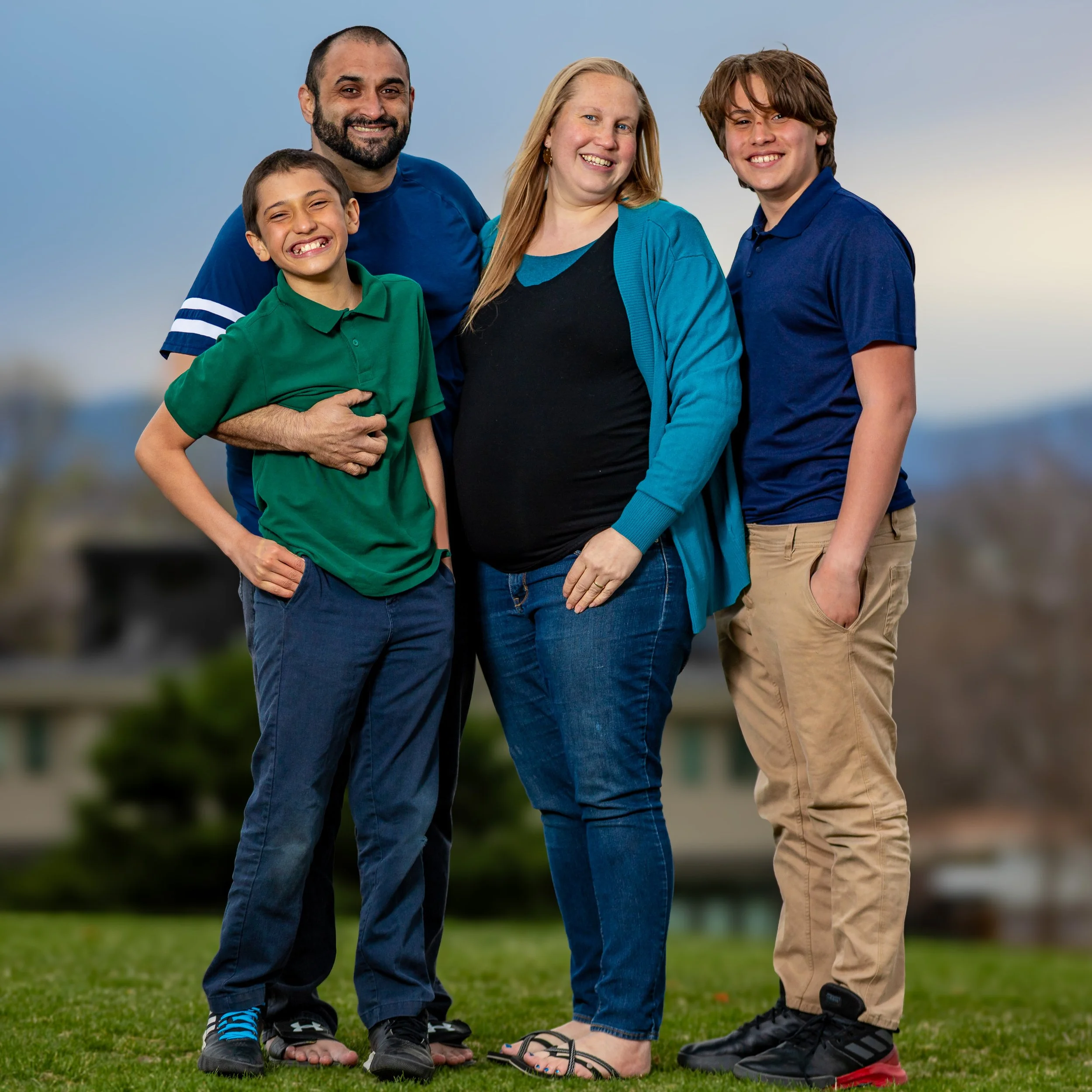 A diverse family of five, including two boys and two adult women, is gathered outdoors on a grassy field under a clear sky. They are all smiling and dressed casually, with some children in polo shirts and the women in jackets and casual shoes.  Famil