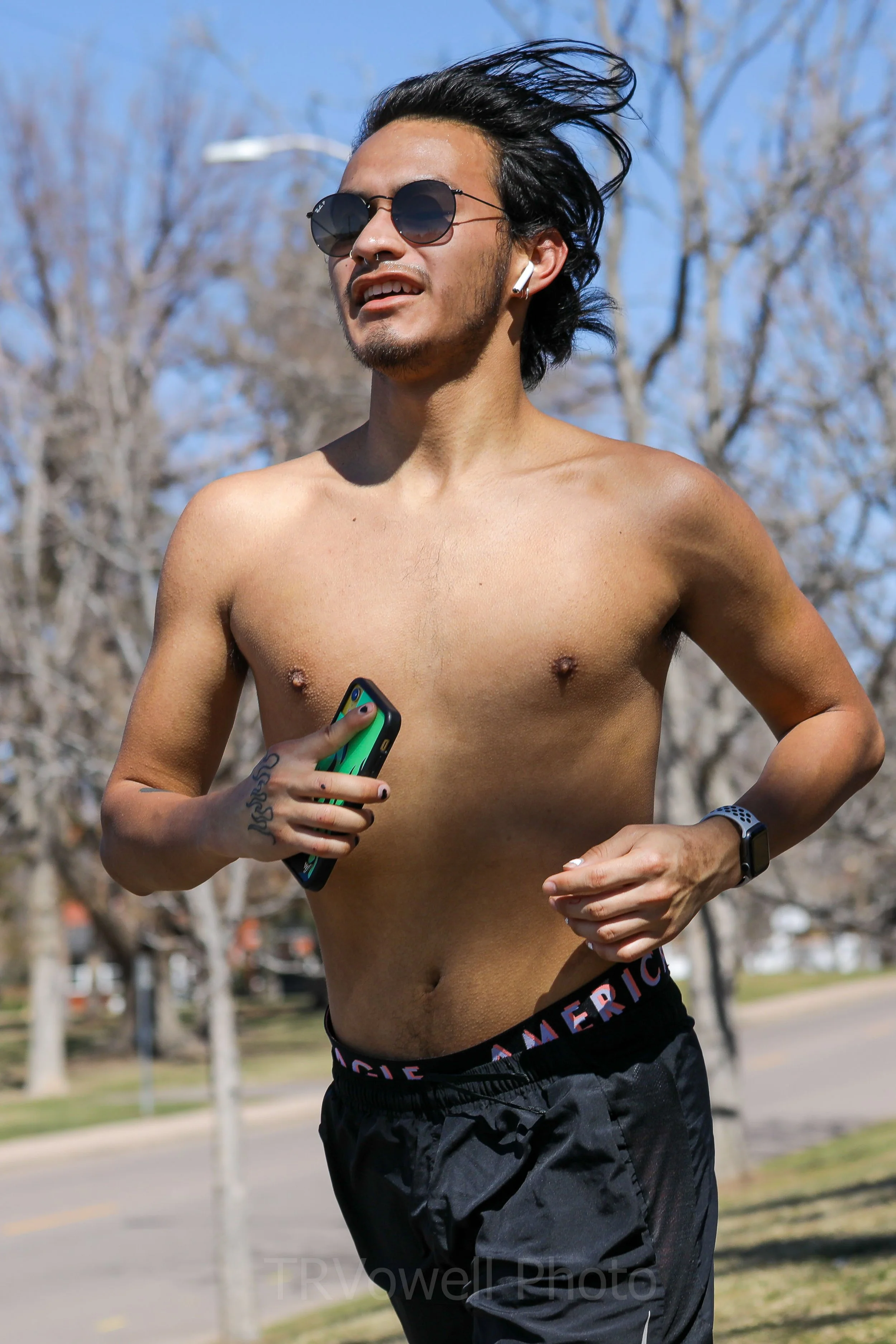 A shirtless man jogging outdoors on a sunny day, wearing sunglasses and a smartwatch, holding a smartphone, with leafless trees in the background.