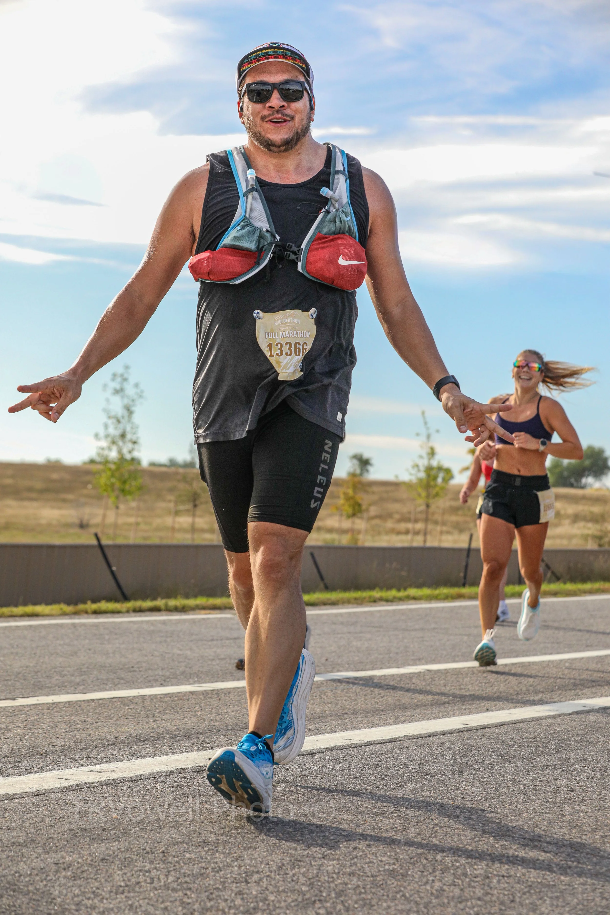A male runner wearing sunglasses, a black sleeveless shirt, black shorts, and blue running shoes, running on a paved road during a marathon, with a female runner in the background.