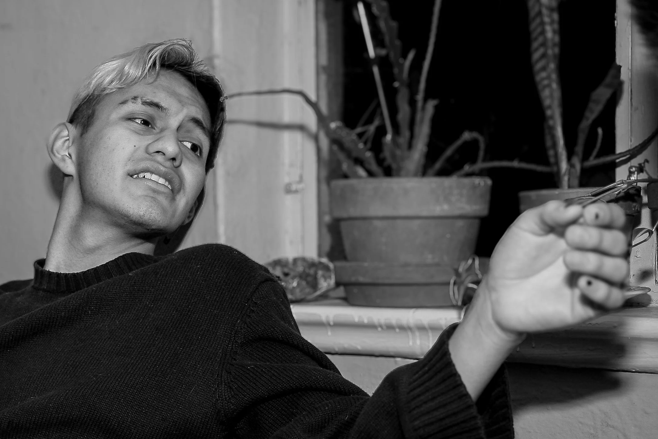 A young man with light-colored hair reclining indoors, with potted plants in the background.