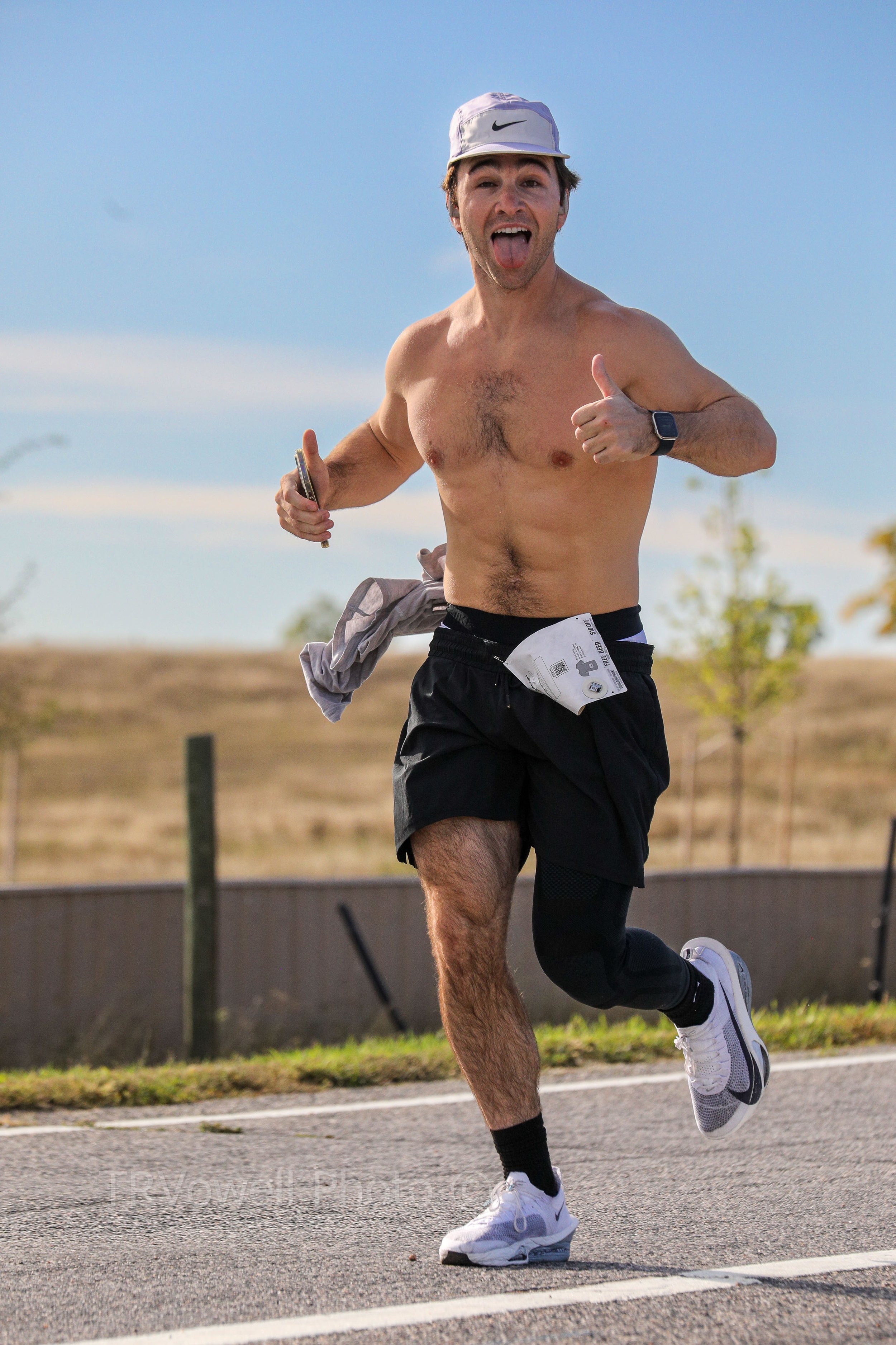 A young man running shirtless on a road, wearing a white cap, black shorts, white running shoes, and a smartwatch, giving a thumbs-up and sticking out his tongue.