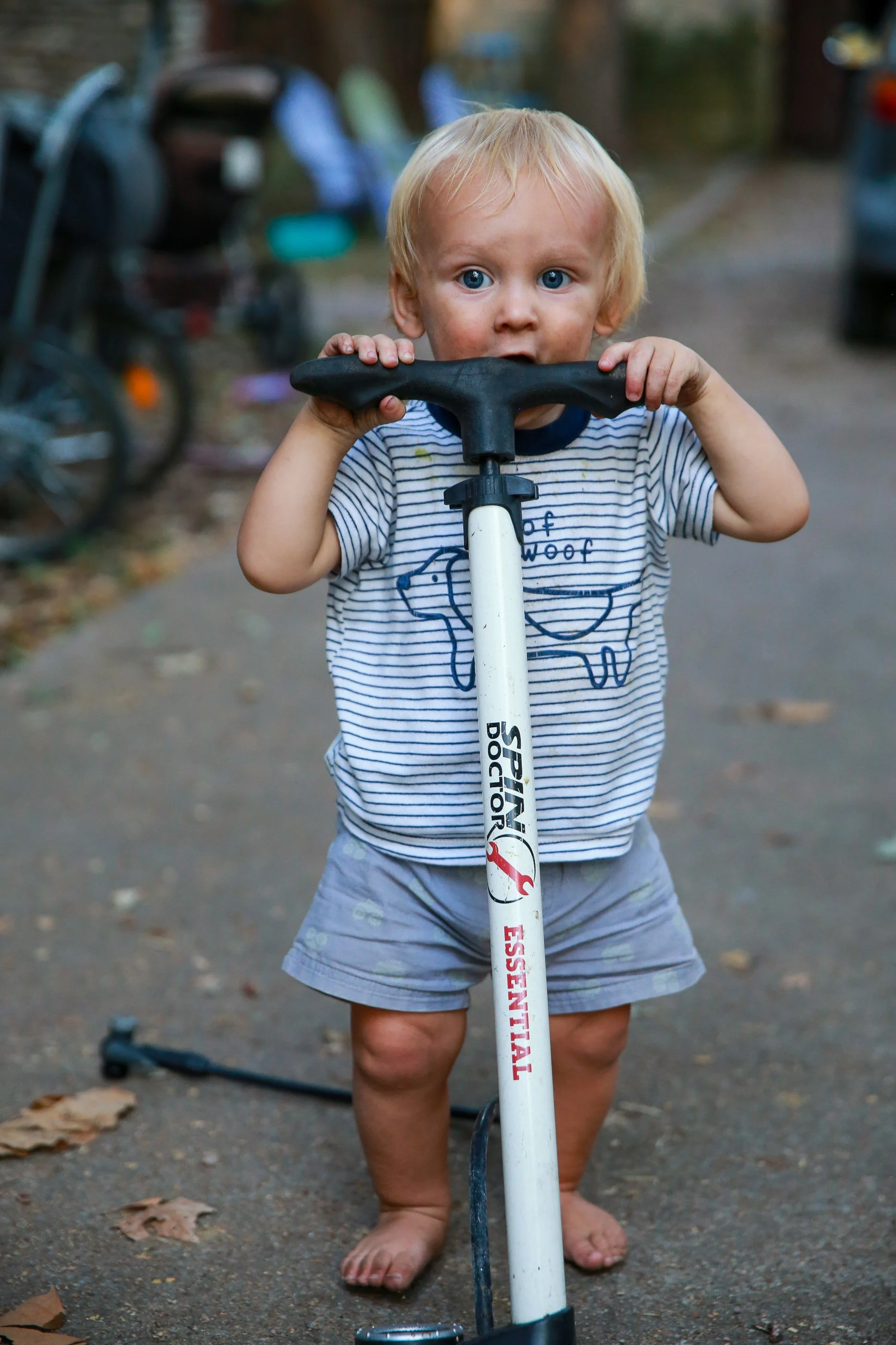 A young blonde-haired boy with blue eyes standing outside, barefoot, holding a scooter handlebar and looking directly at the camera. He is wearing a striped t-shirt and shorts, with a curious expression. Blurred trees, a parked bicycle, and some cars