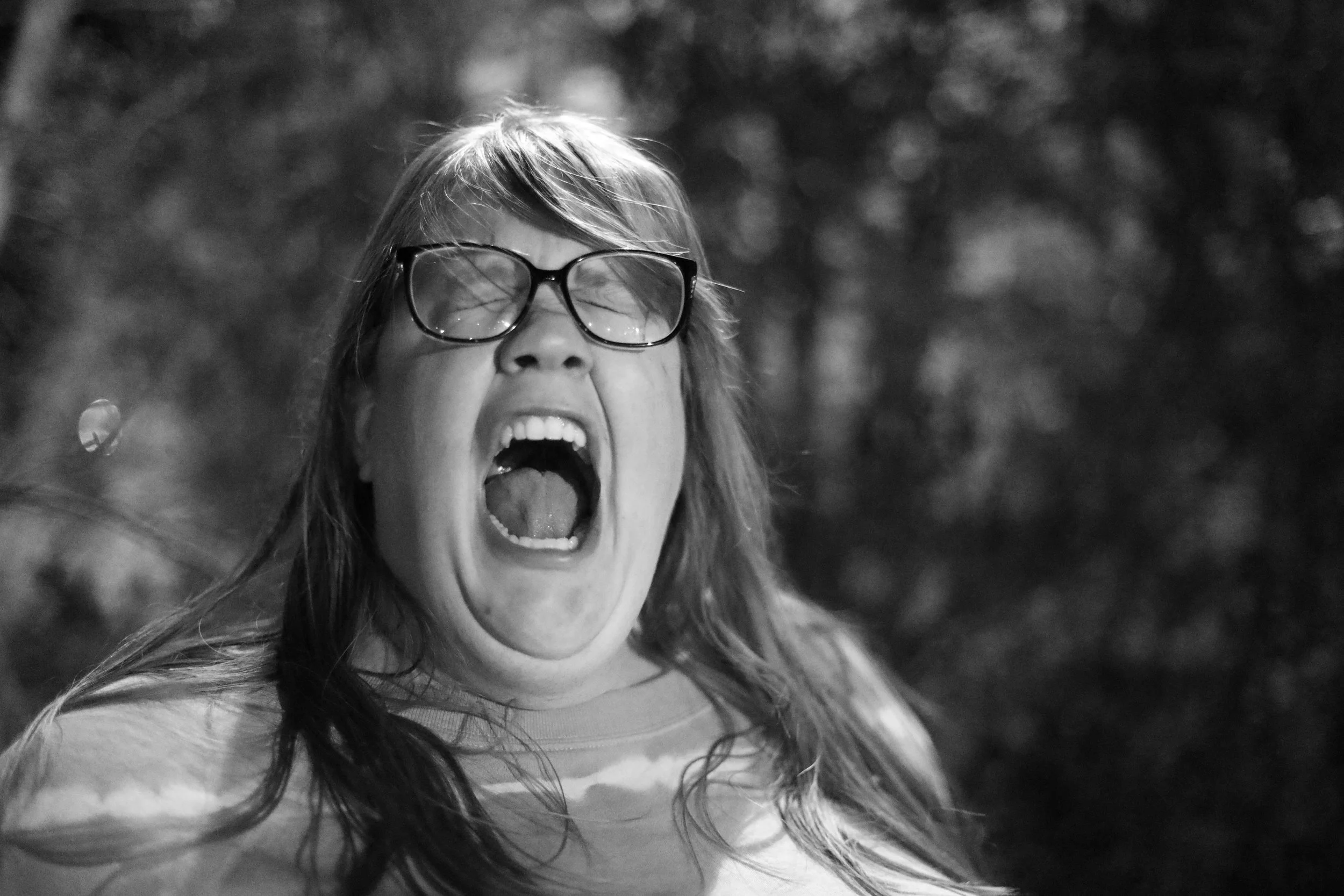 A woman with glasses and long hair appears to be yelling or screaming with her mouth wide open, outdoors with blurred trees in the background. Real emotions real life documentary editorial style photography and portraits