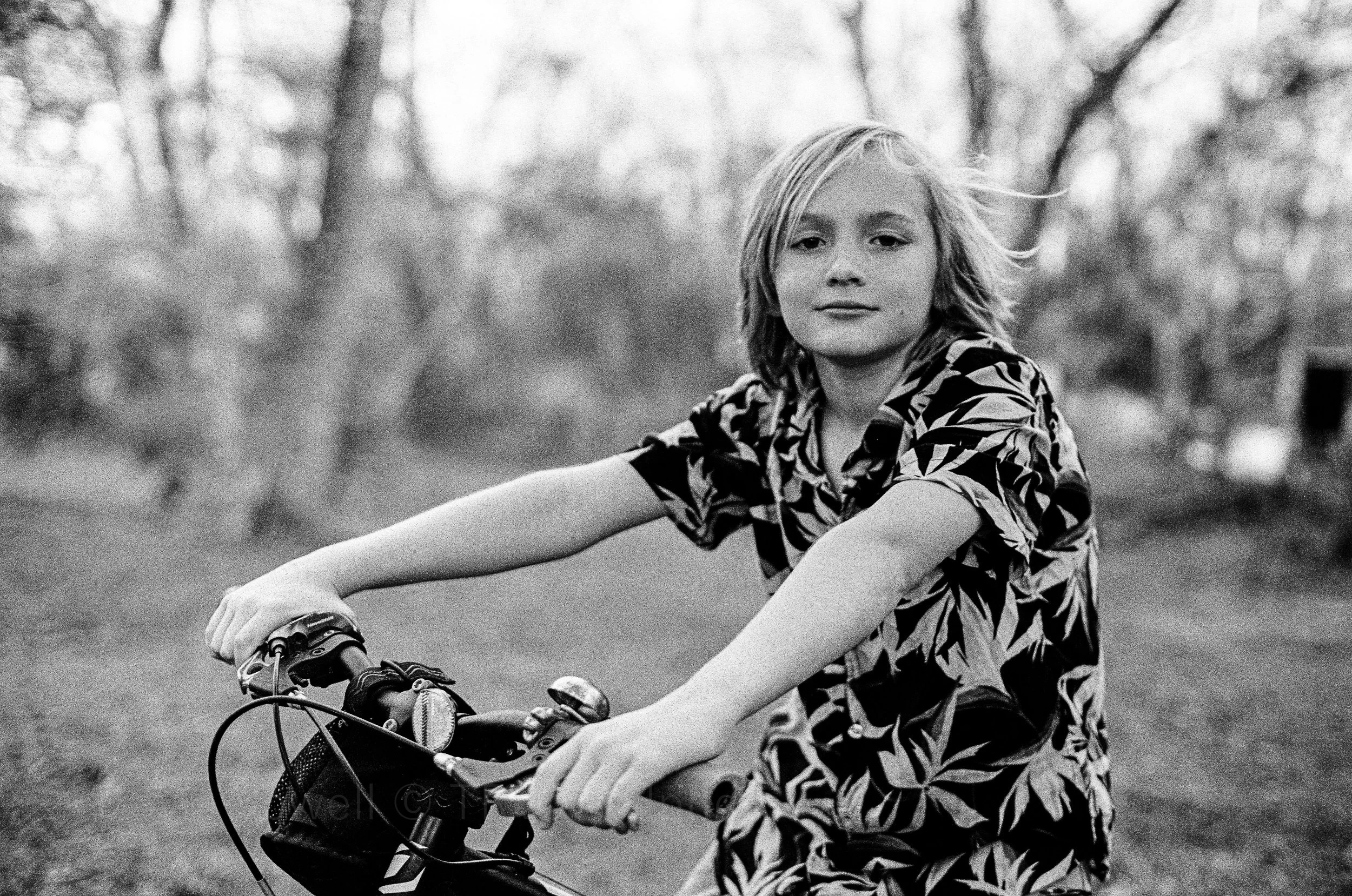 portrait photography of A young child with short hair wearing a patterned shirt riding a bicycle outdoors in a park or forest setting, with trees in the background.  Classic traditional old school retro Black and White B&W BW 35mm film photography sh