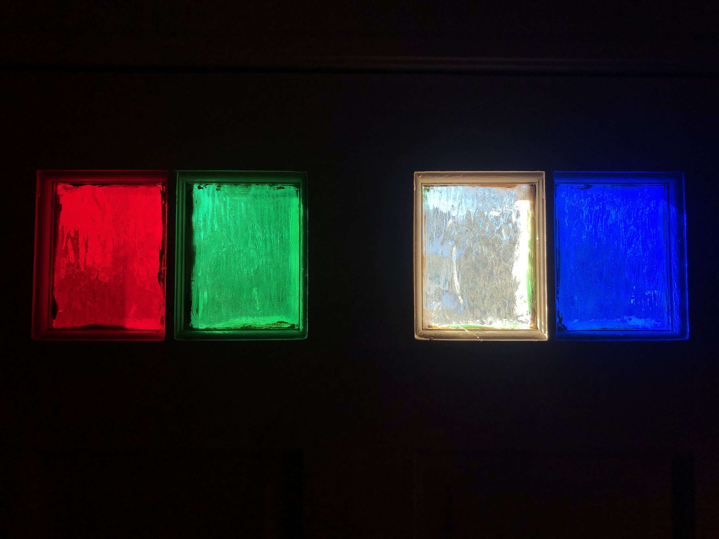 Four small stained glass windows in a dark wall, from left to right: red, green, clear, and blue.