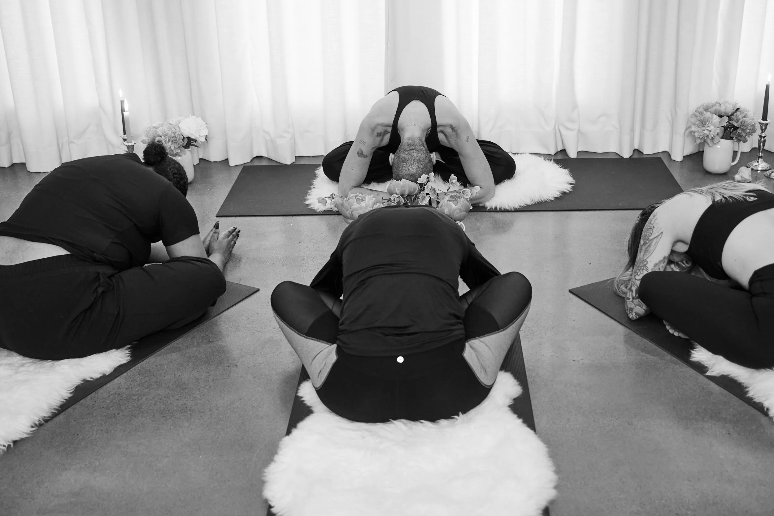Four people participating in a yoga or meditation class, seated on mats with fluffy cushions, in a room with white curtains and candles, and floral decorations.