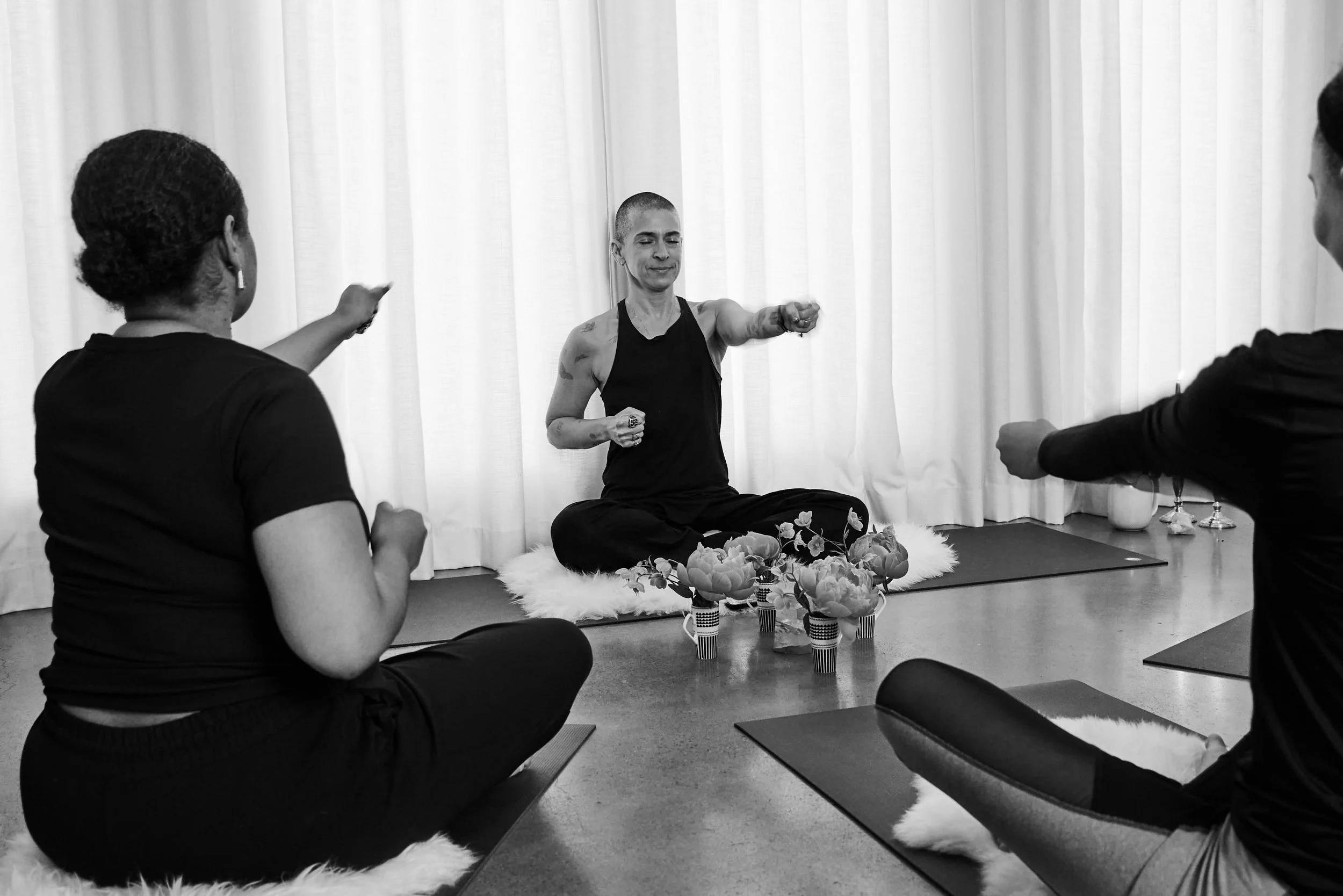 Three people participating in a meditation session, sitting cross-legged on cushions in a room with curtains, with Sam in the center leading the practice.