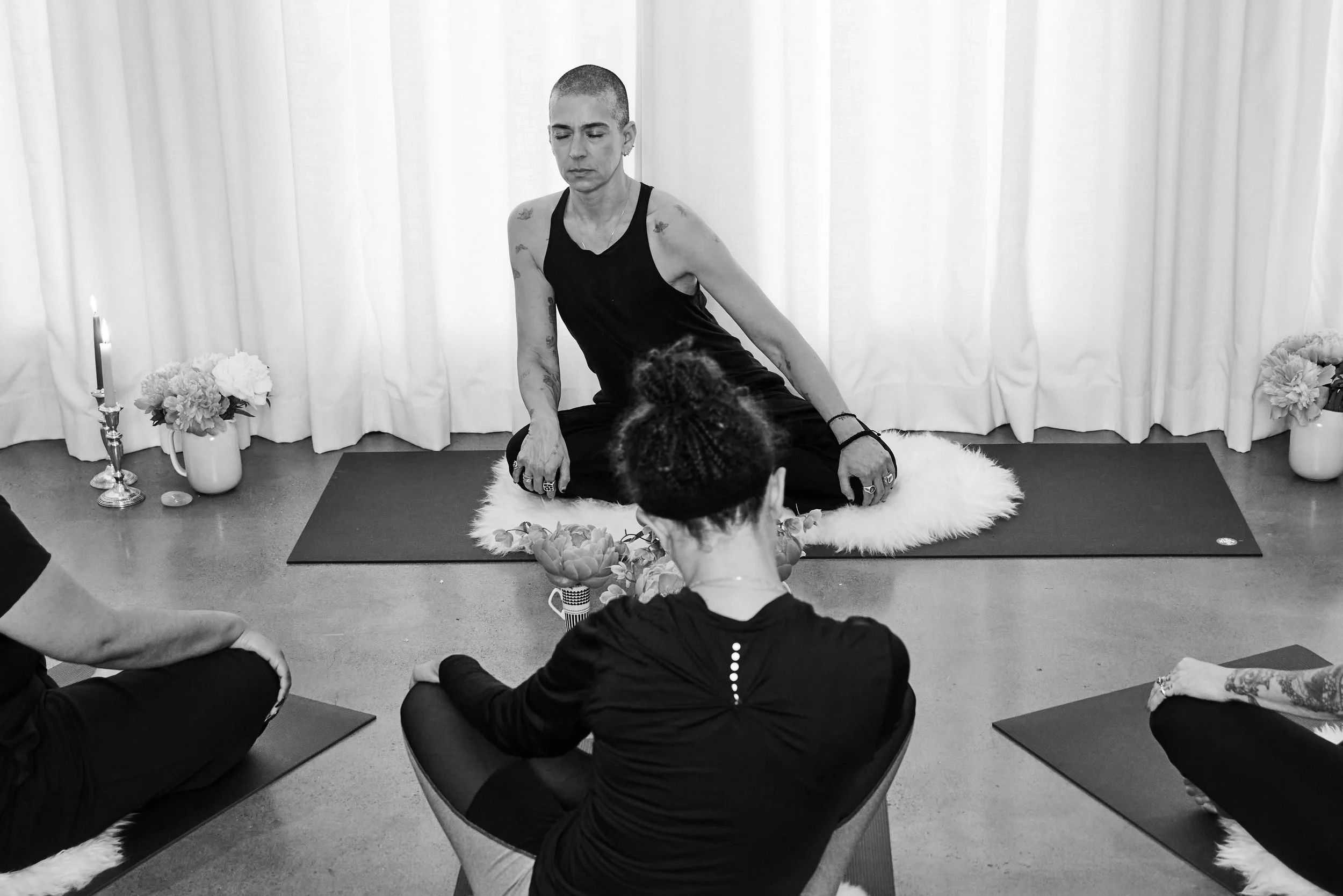 Sam sitting cross-legged on a fluffy rug, leading a meditation / yoga session with group members sitting on mats in a room with white curtains, flowers, and candles.
