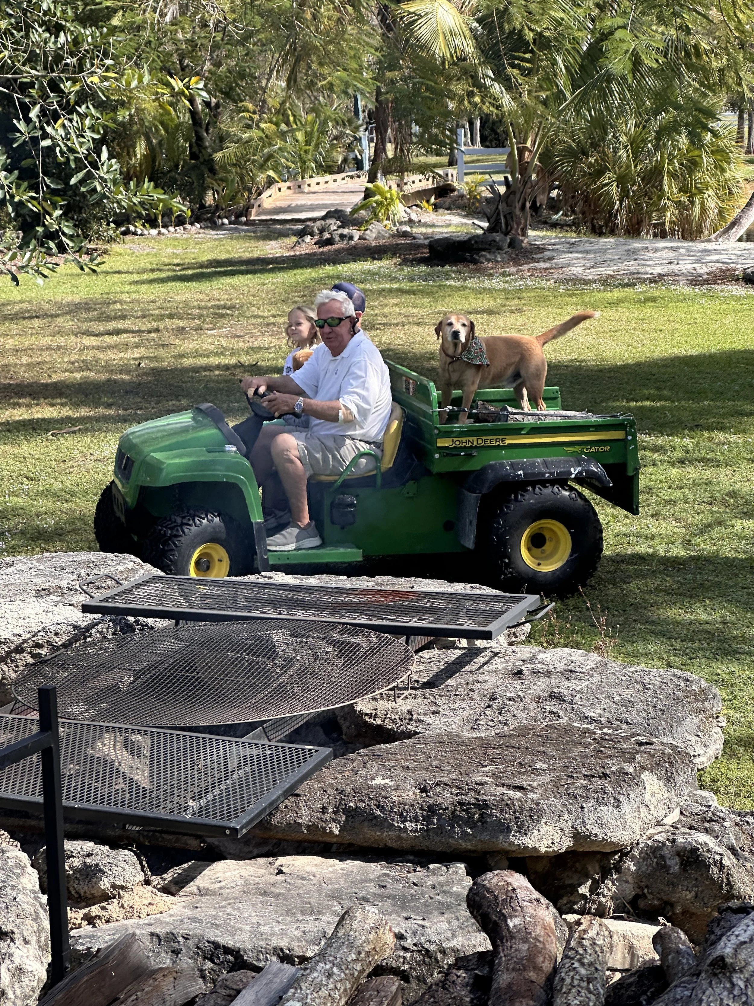 Man and girl riding a small green John Deere Gator utility vehicle in a park with palm trees, and a dog sitting in the cargo bed wearing a bandana. Barr Farms Family Farm in Florida