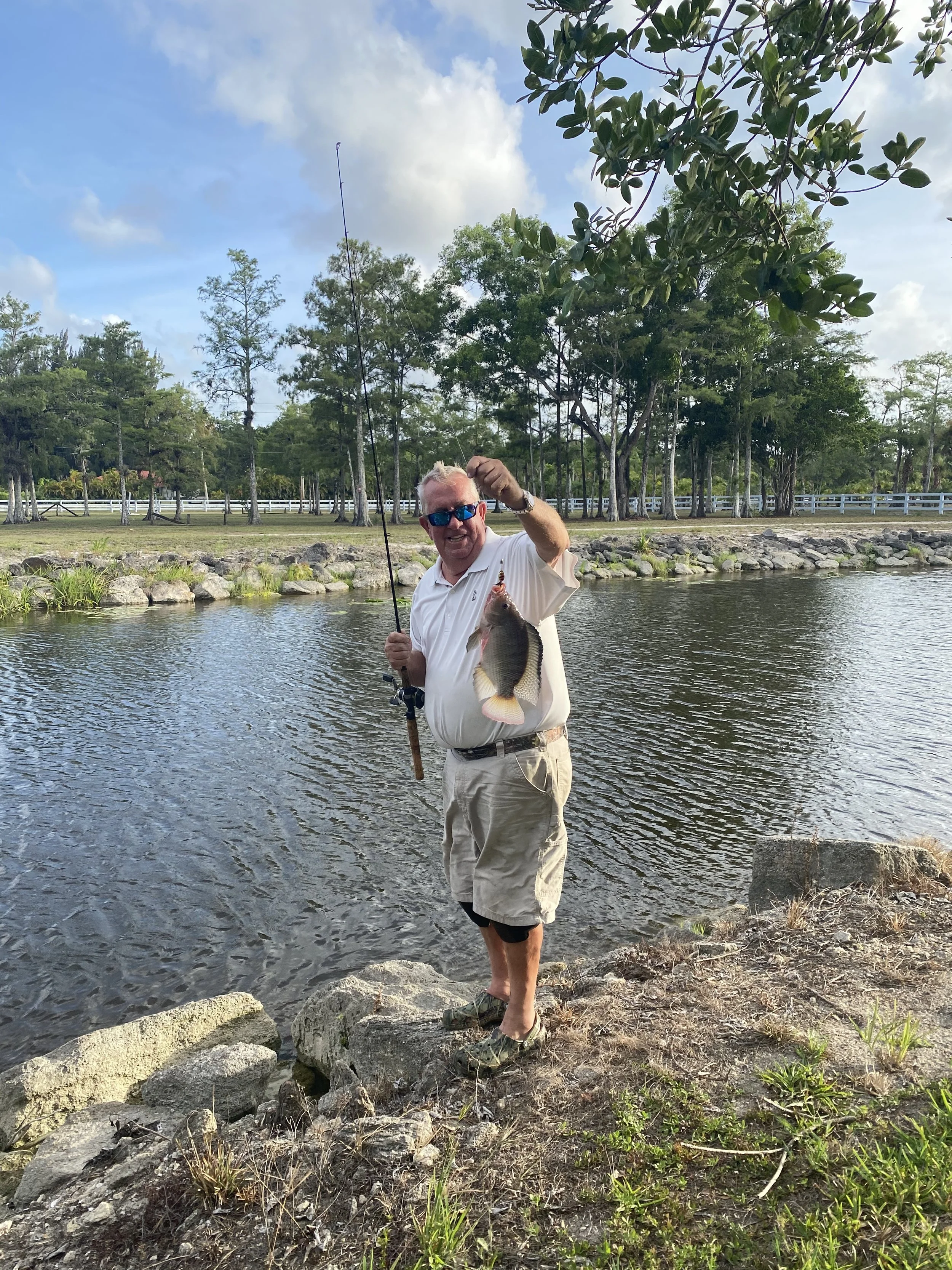 A man standing on rocks by a pond, holding a fishing rod and a caught fish, smiling, with trees and a fence in the background.