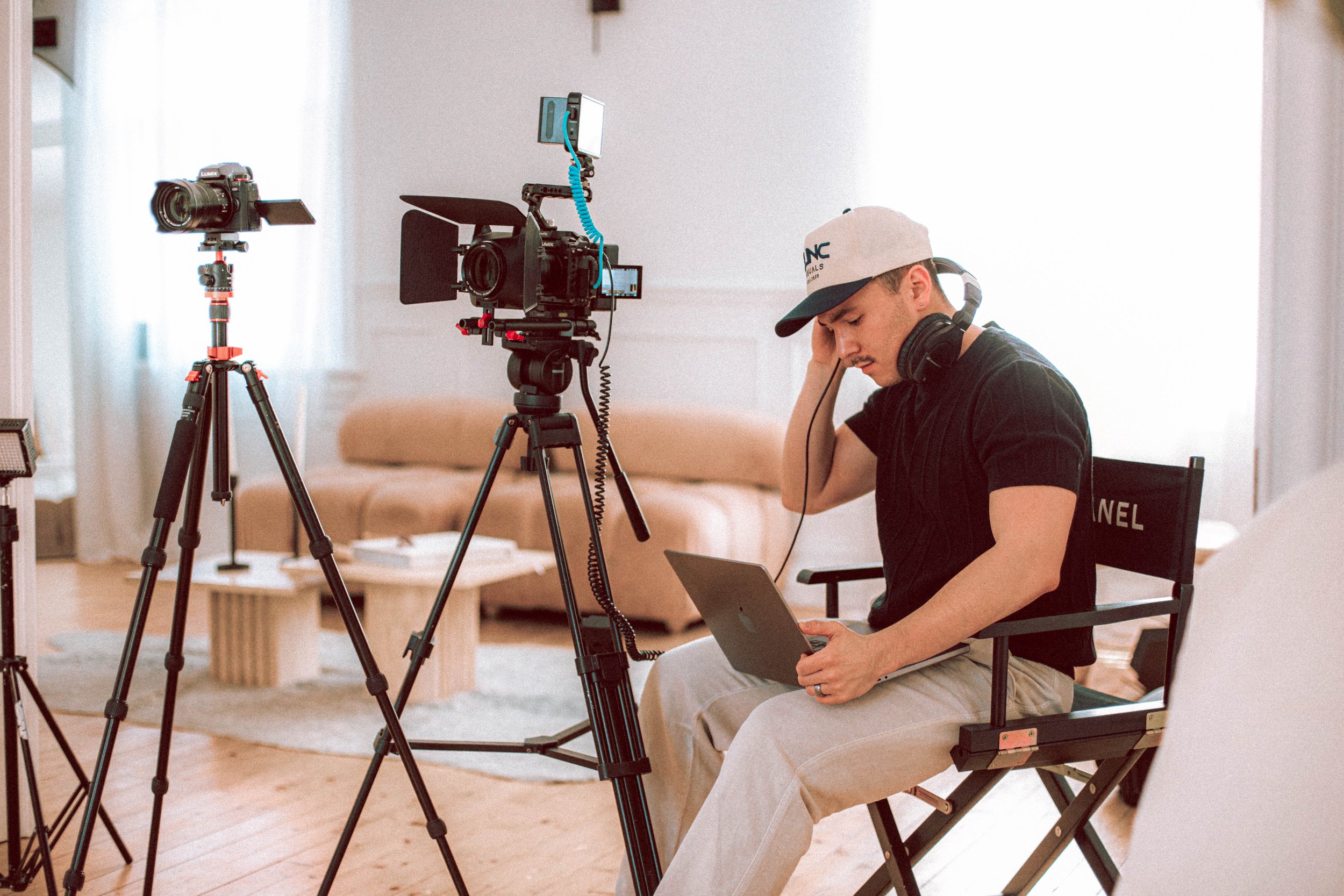 Young man operating professional film camera equipment in a bright, modern living room, wearing headphones and a cap, sitting on a director's chair, with additional cameras on tripods around him.