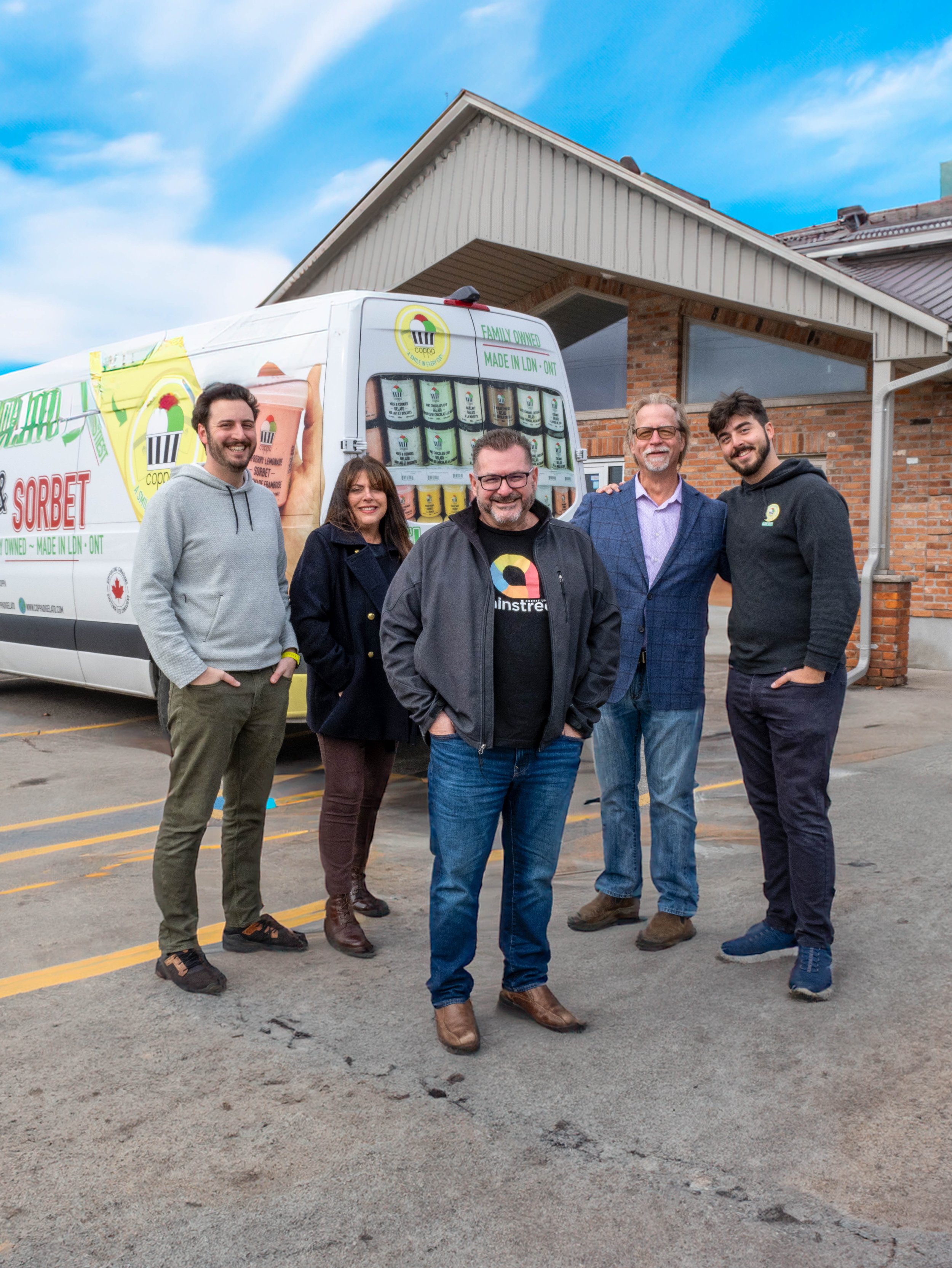 Group of five people standing outdoors in front of a branded ice cream truck with a partly cloudy sky above and a brick building behind.
