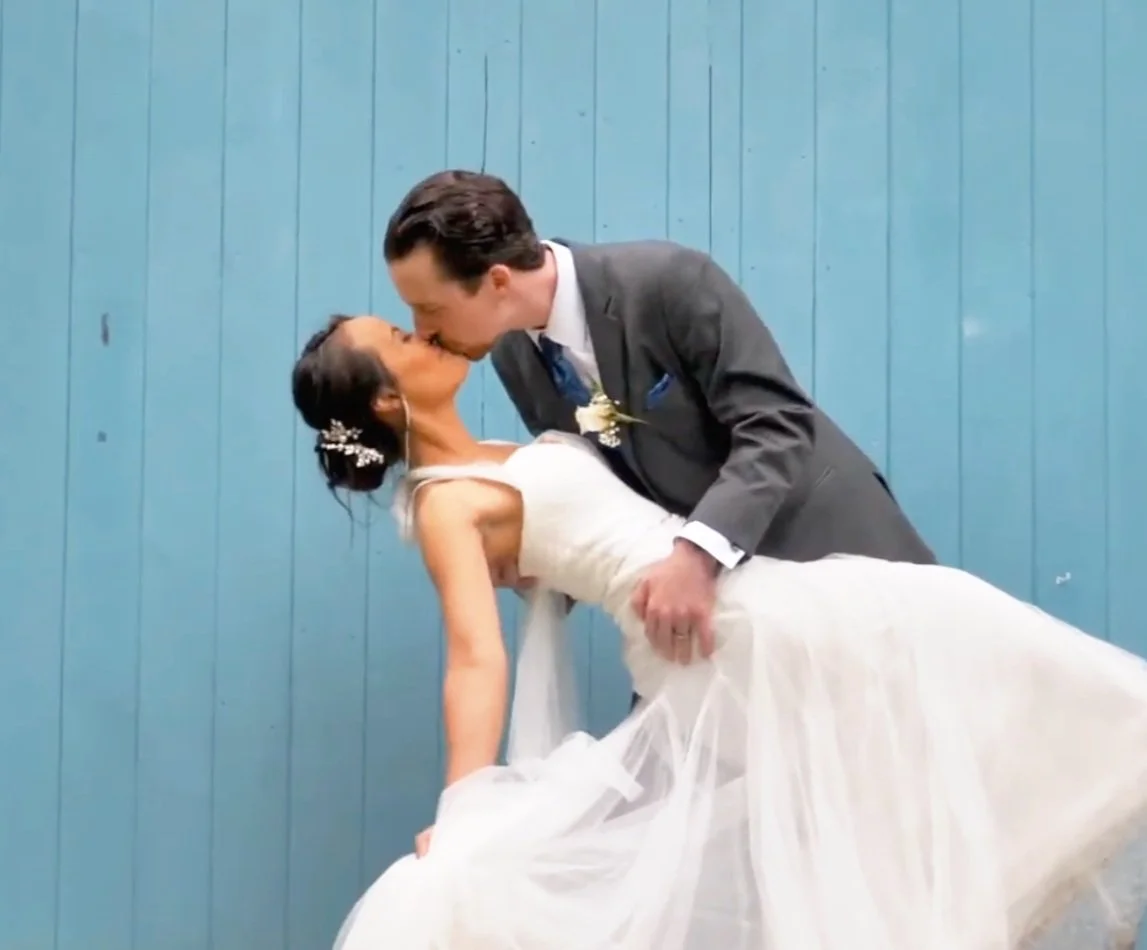 A man in a gray suit dips a woman in a white wedding dress for a kiss against a blue wooden wall.