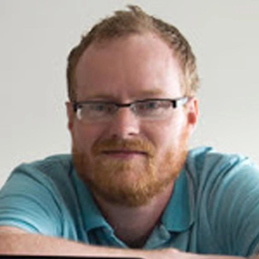 A man with red hair and a beard wearing glasses and a blue shirt, smiling, with a light-colored background.