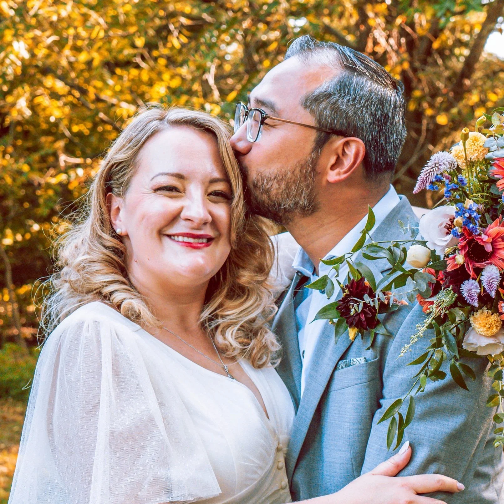 A happy woman in a white dress and a man in a gray suit share a kiss on the cheek during a wedding or special event outdoors, with a colorful floral bouquet and autumn trees in the background.