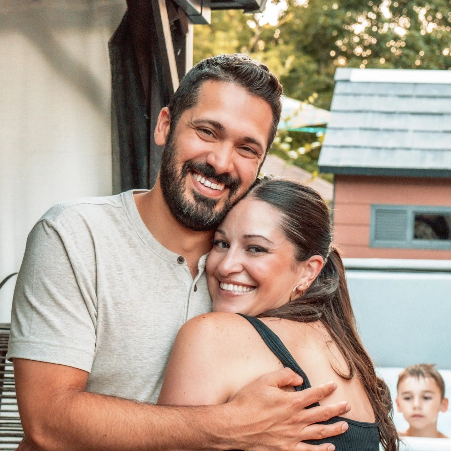 A smiling man and woman embracing outdoors, with a child in the background.