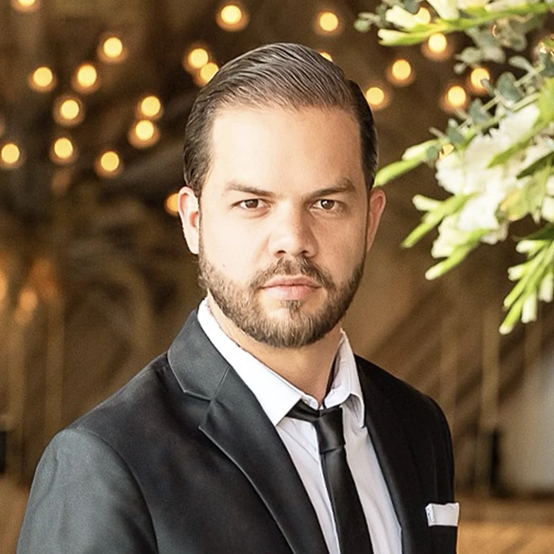 A man with a beard in a black tuxedo, white shirt, and black tie, posing indoors at a formal event with warm lighting and floral decorations.