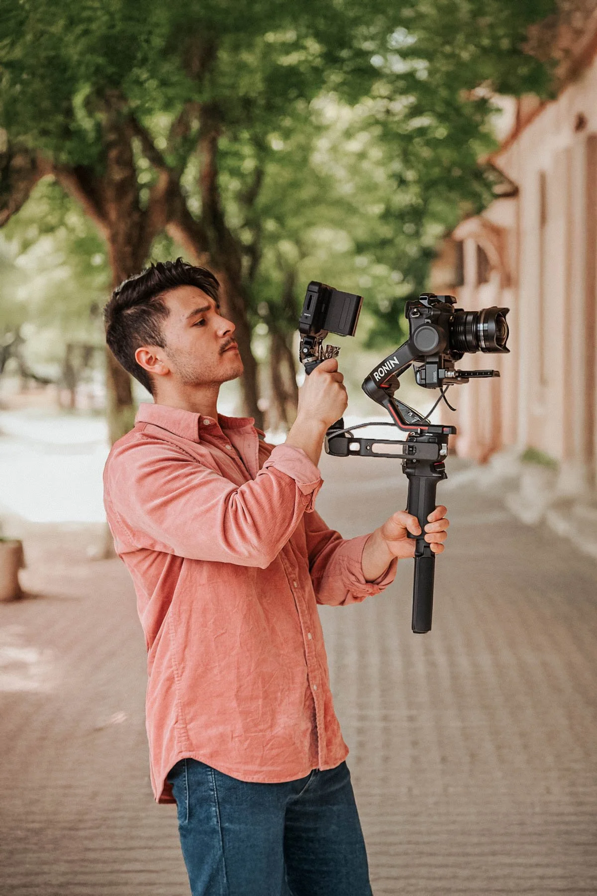 A man holding a camera stabilizer with a mounted camera, filming or setting up a shot outdoors on a tree-lined street.