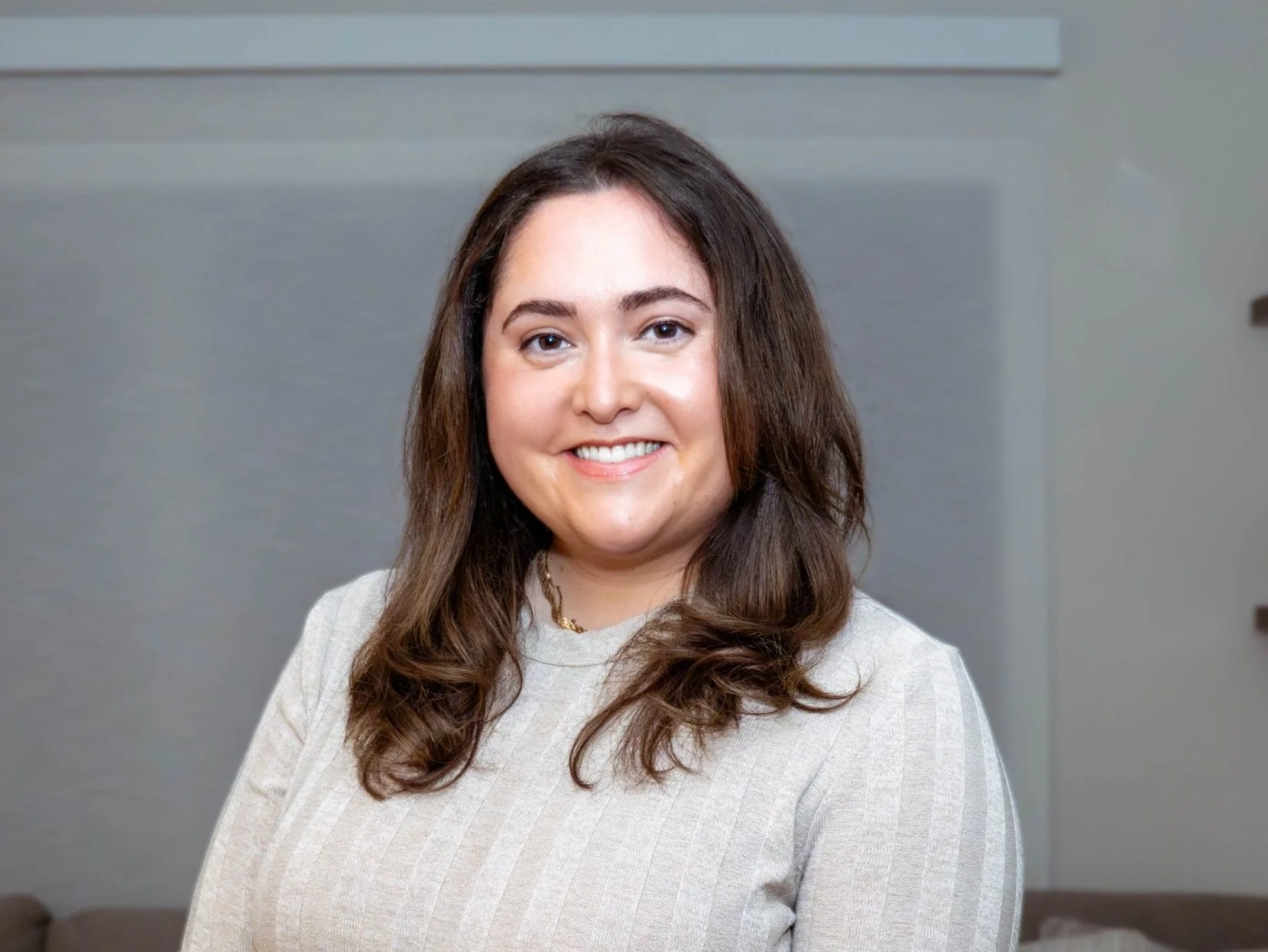 A smiling woman with shoulder-length brown hair, wearing a light-colored striped top, standing in a modern indoor environment.