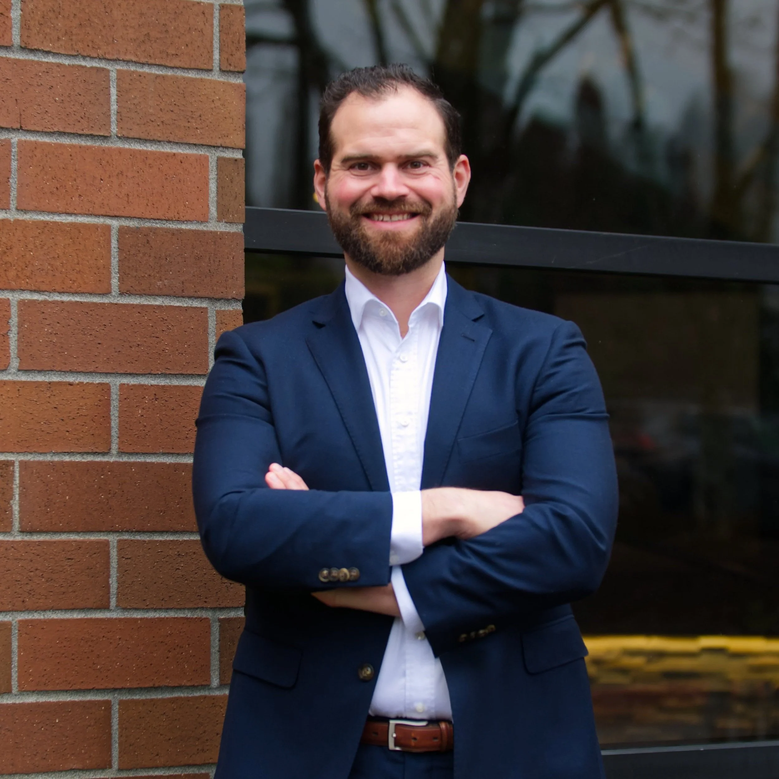 A man with a beard and short dark hair standing outside near a brick wall, smiling with arms crossed, wearing a navy blazer and white shirt.
