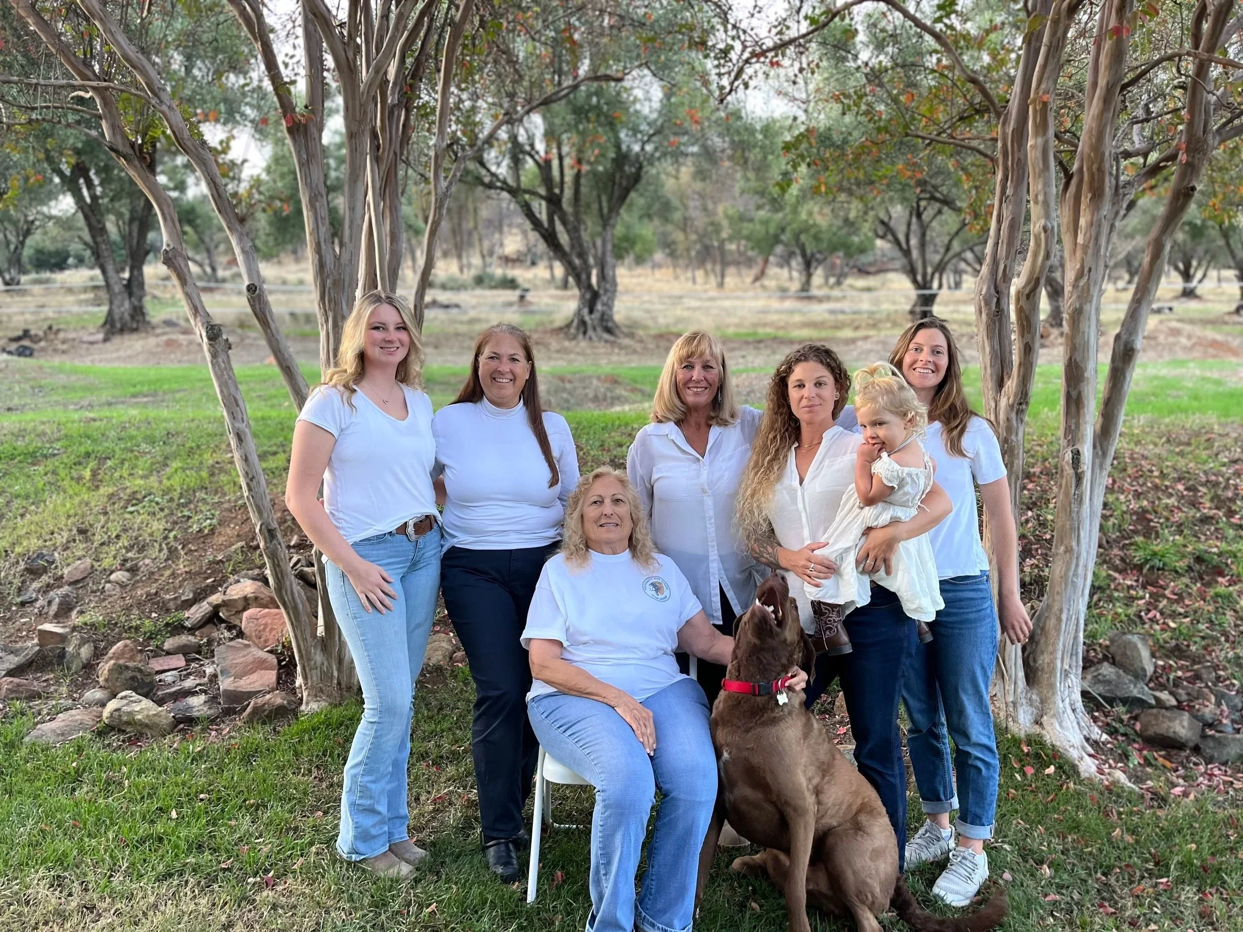 A group of eight women, one child, and a large brown dog posing outdoors under trees with autumn leaves. They are smiling and facing the camera.