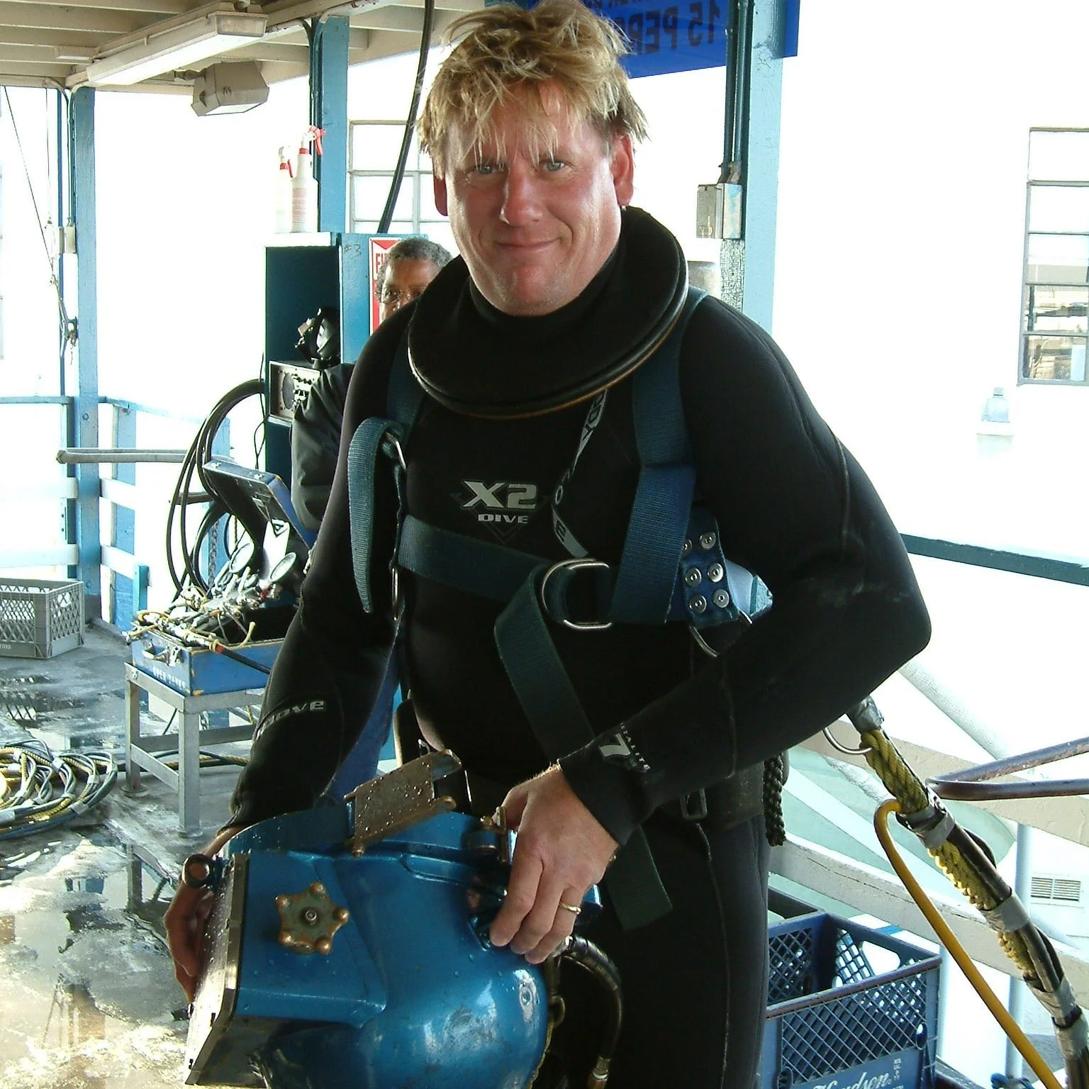 A man in a scuba diving suit holding a blue diving helmet inside a boat or dock area.