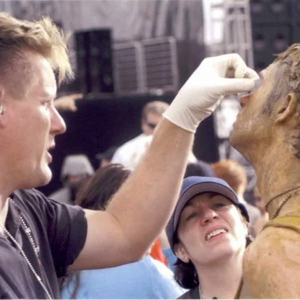 Makeup artist applying special effects makeup to a person in a costume, with a crowd in the background.