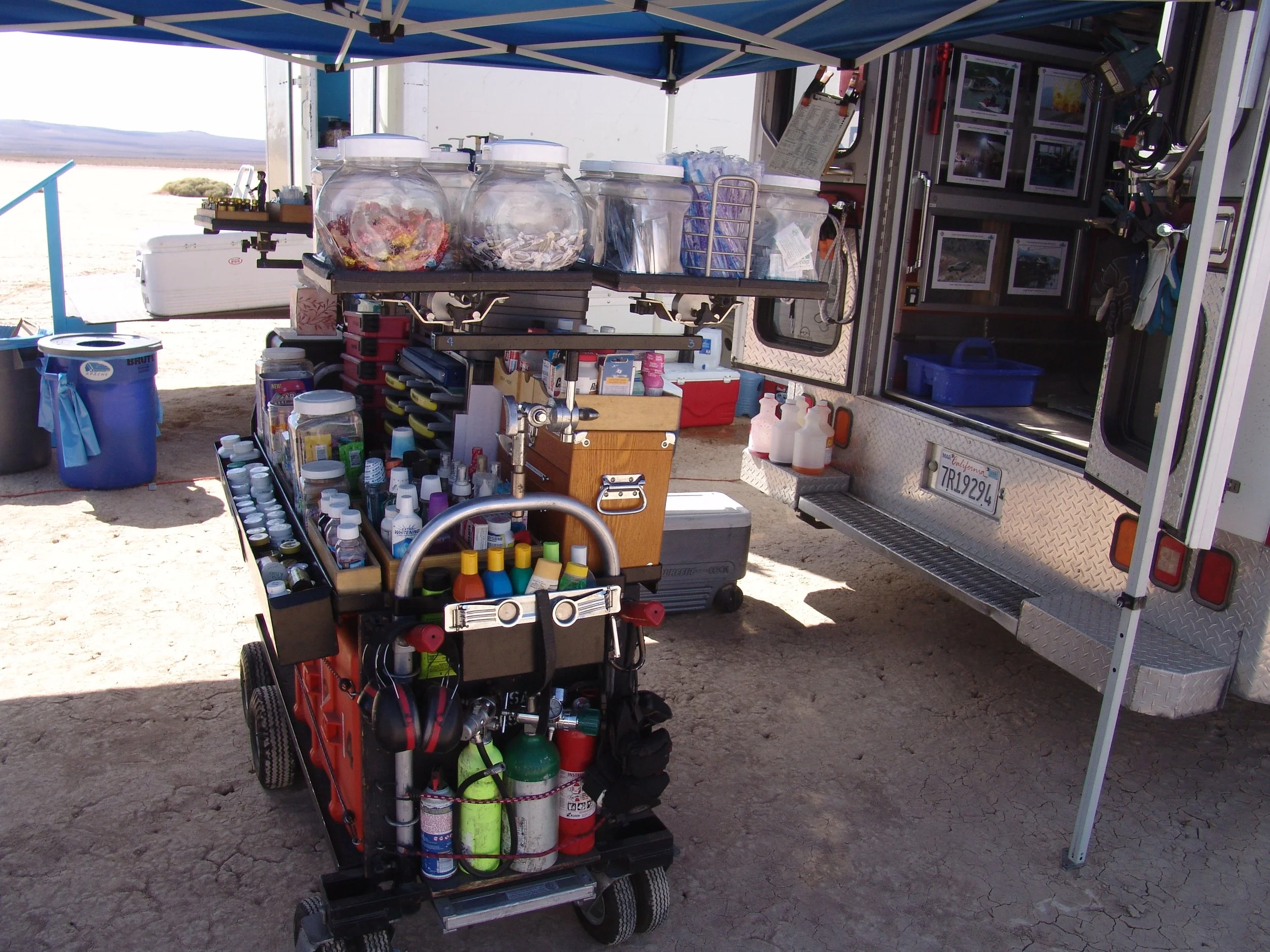 Arrangement of supplies and equipment at a mobile water testing station, including bottles, containers, tools, and a mobile cart with dive gear, set up outdoors with a canopy.