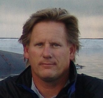 A man with light brown hair and fair skin standing outdoors near a body of water.