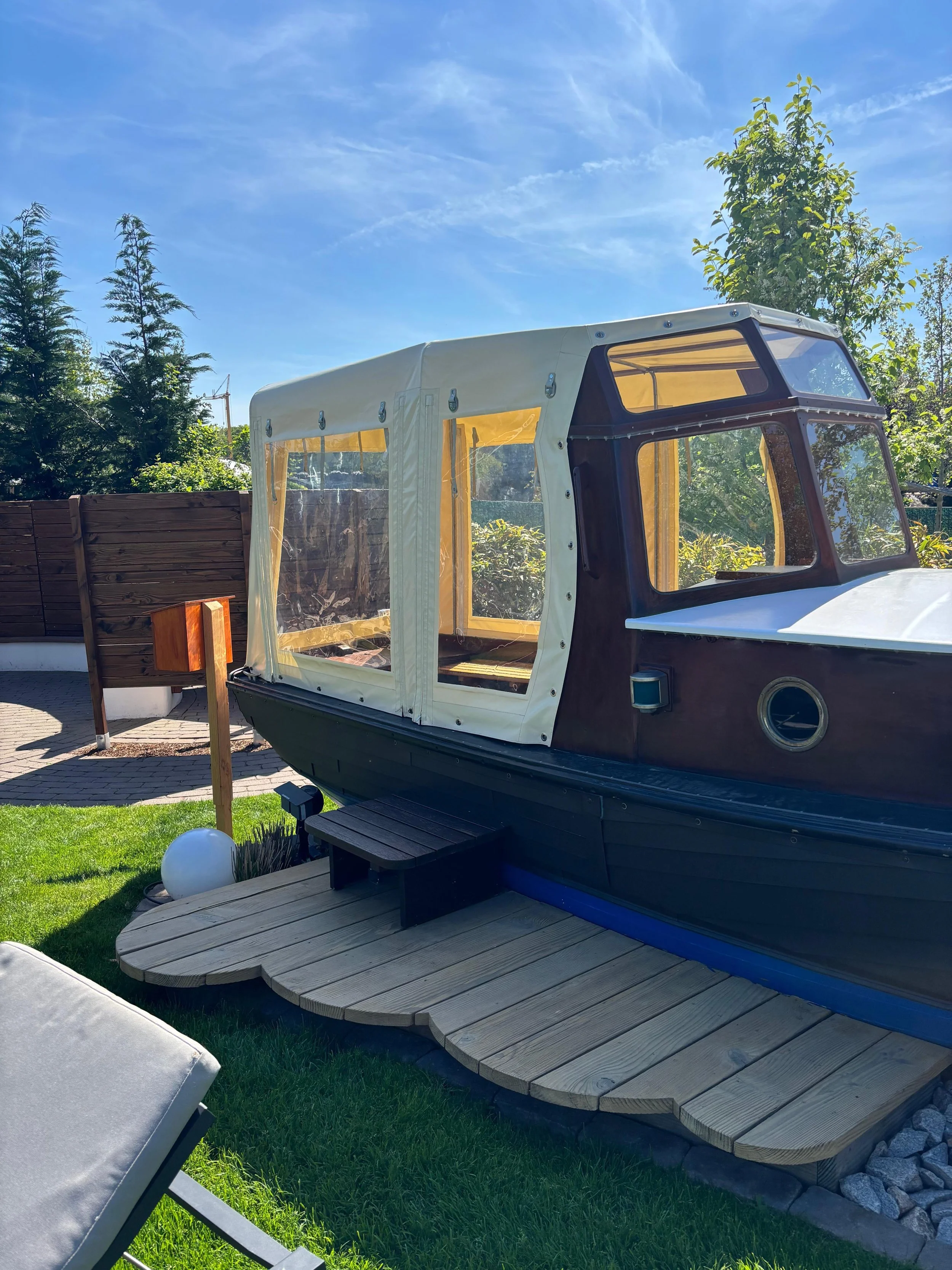 Ein kleines Boot mit einem hellen Verdeck auf einem Holzsteg vor einem Garten mit Gras, Bäumen und einem blauen Himmel.