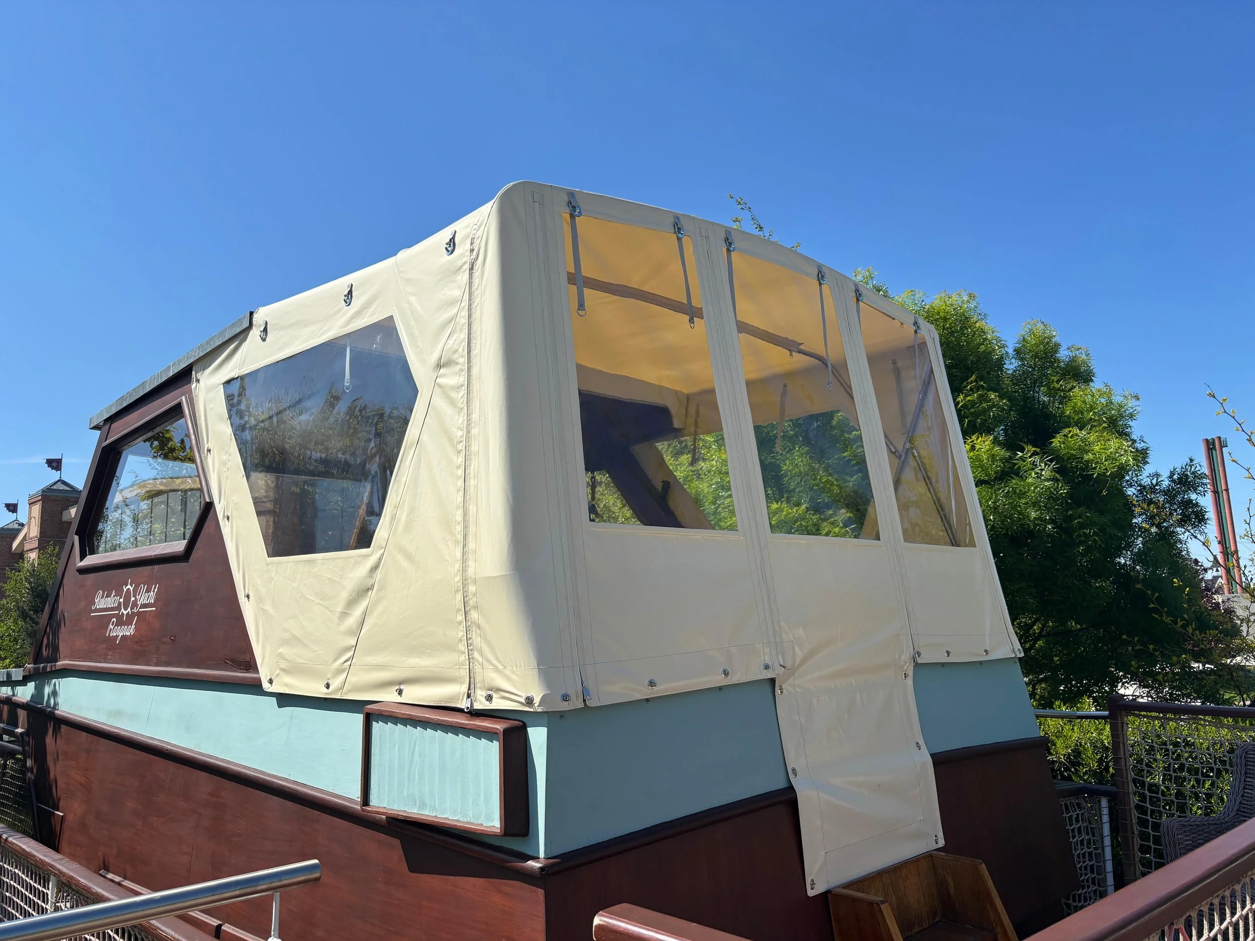 Ein Hausboot mit Holzverkleidung und einer beige Verkaufsplane im Sonnenschein vor blauem Himmel, umgeben von grünen Bäumen.