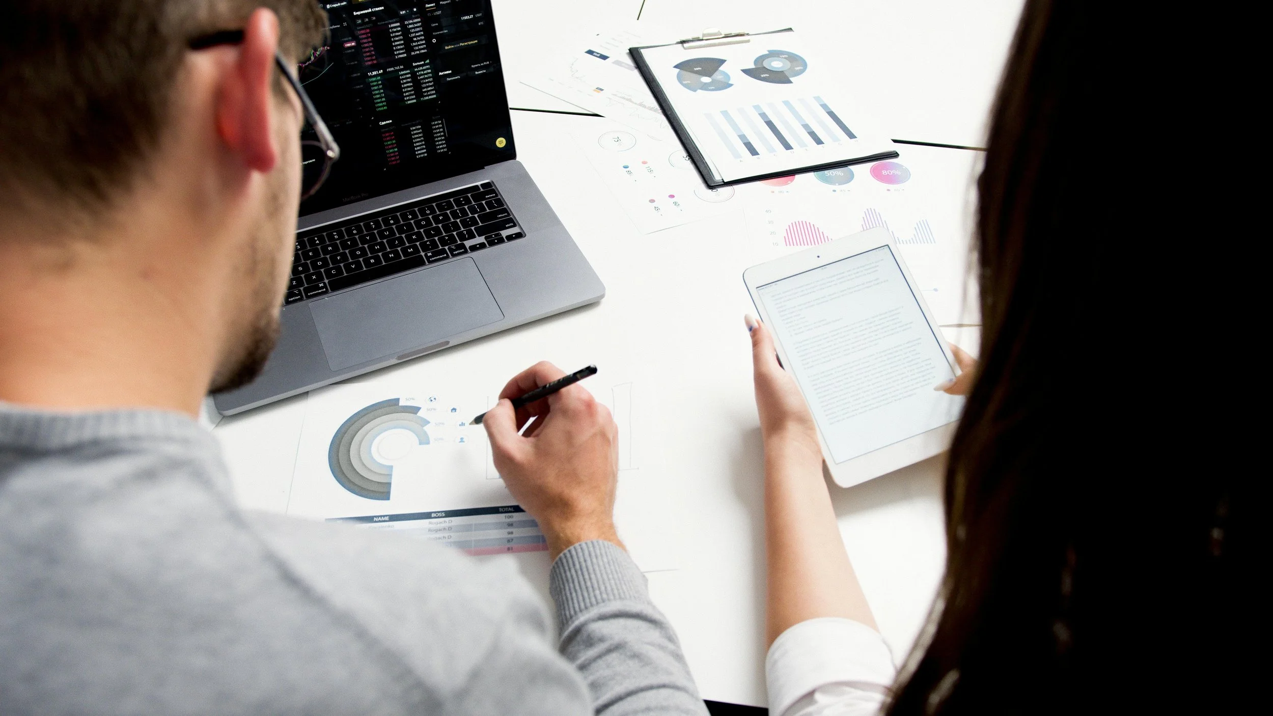 Two people collaborating at a desk with a laptop, tablet, and printed charts.