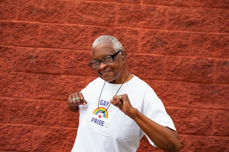 A Black woman smiles and flexes her arms in a show of strength, wearing a white Gay Pride t-shirt with a rainbow graphic, standing in front of a red brick wall
