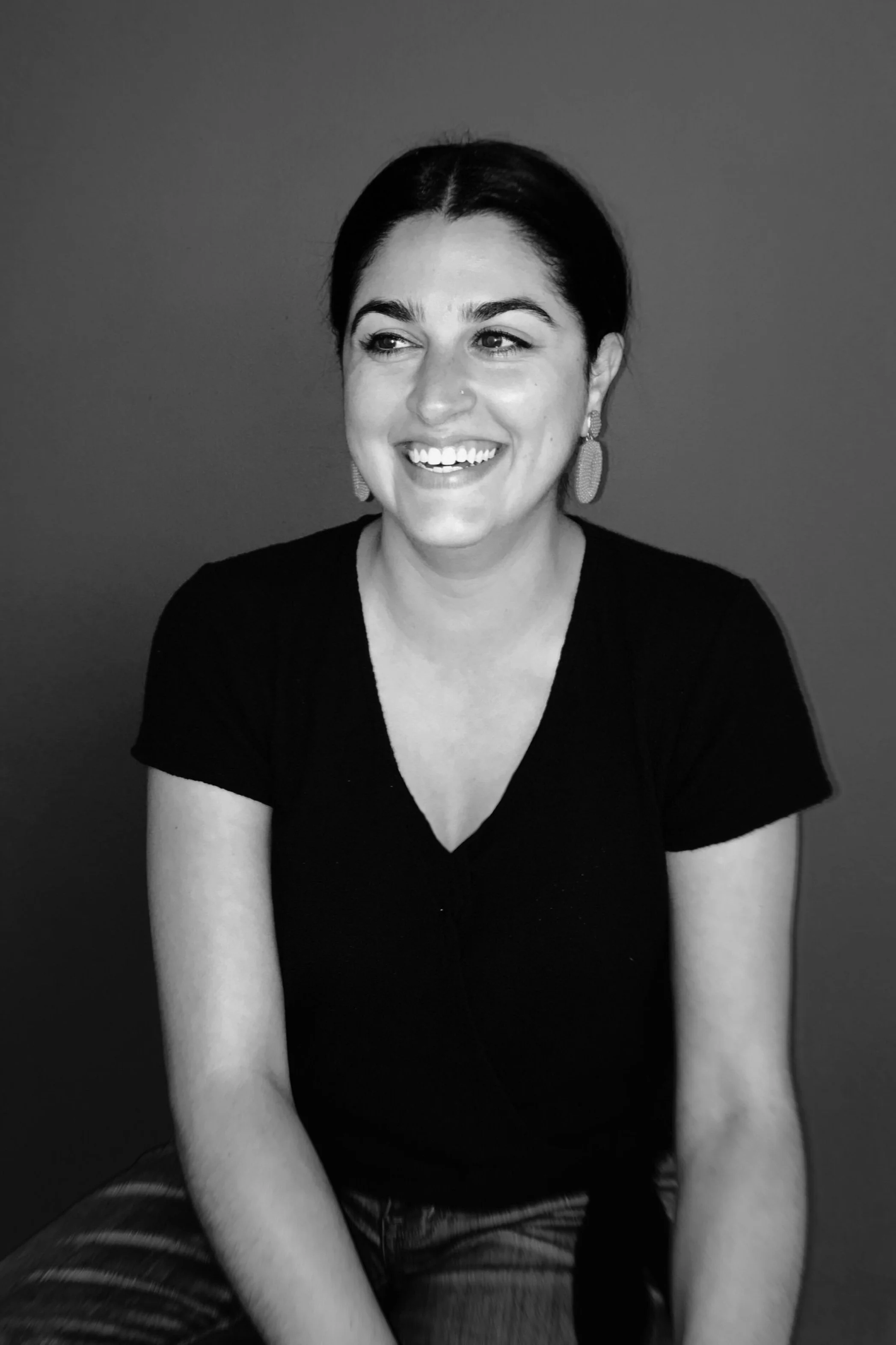 Black and white portrait of a smiling woman with dark hair, earrings, and a black shirt, sitting against a plain background.