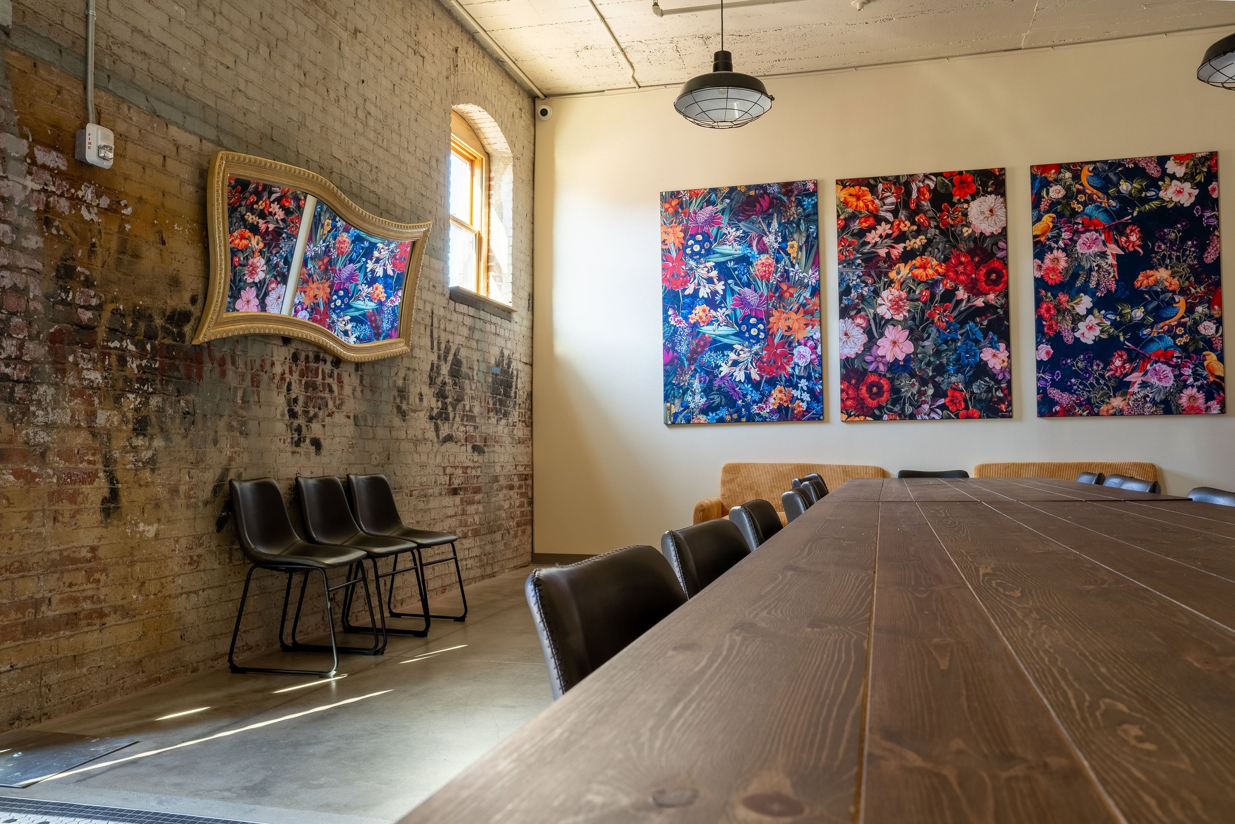 A conference room with a long wooden table, black chairs, a brick wall with three colorful floral paintings, and a mirror reflecting part of the room.