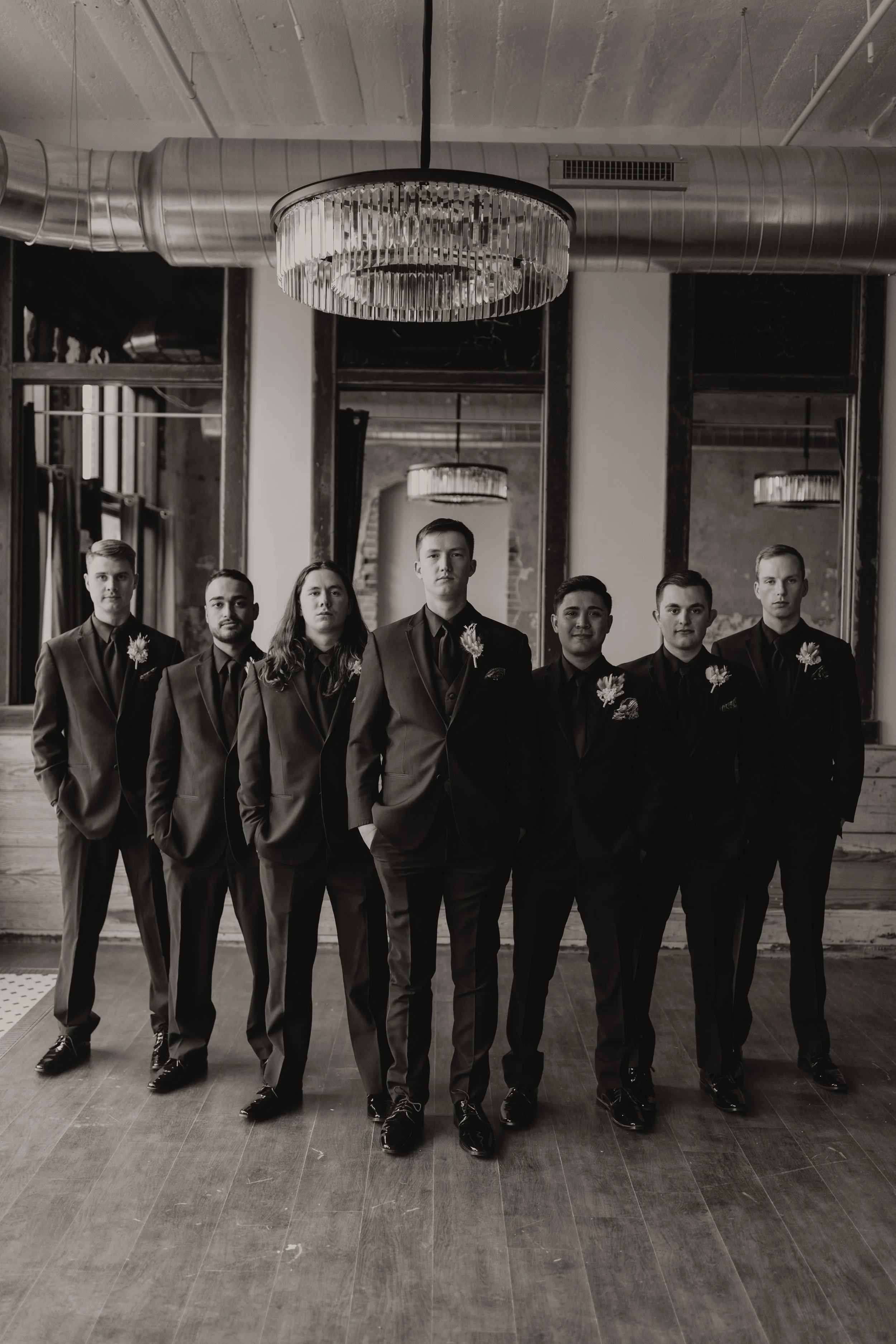 Black-and-white photo of seven men in tuxedos standing in a line indoors.