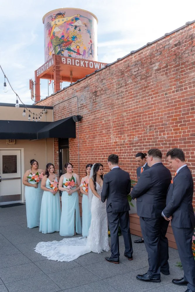 A wedding ceremony taking place outdoors against a brick wall, with the bride and groom exchanging vows and bridesmaids and groomsmen standing nearby.