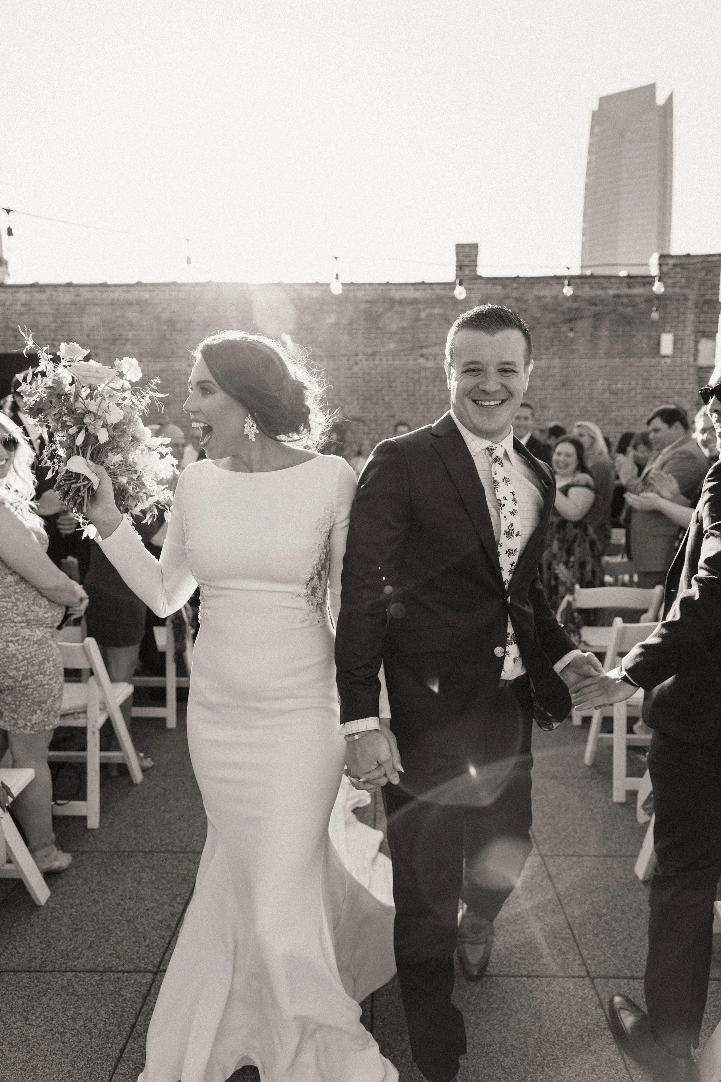 A bride and groom holding hands and smiling at their wedding reception on a rooftop, with guests clapping and a cityscape in the background.