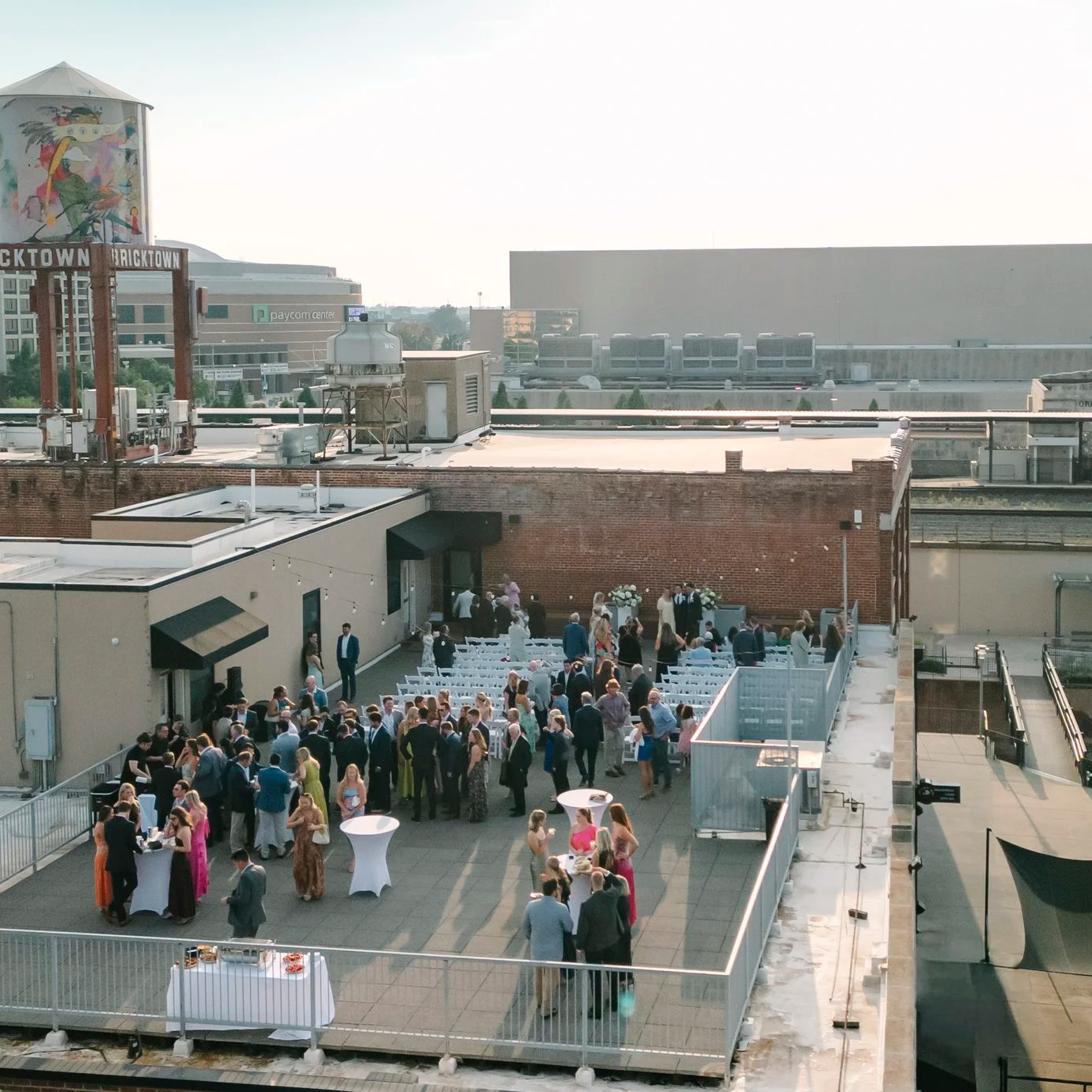 Outdoor wedding ceremony with guests seated on white chairs facing a floral arch on a brick wall, on a rooftop with string lights and city buildings in the background.