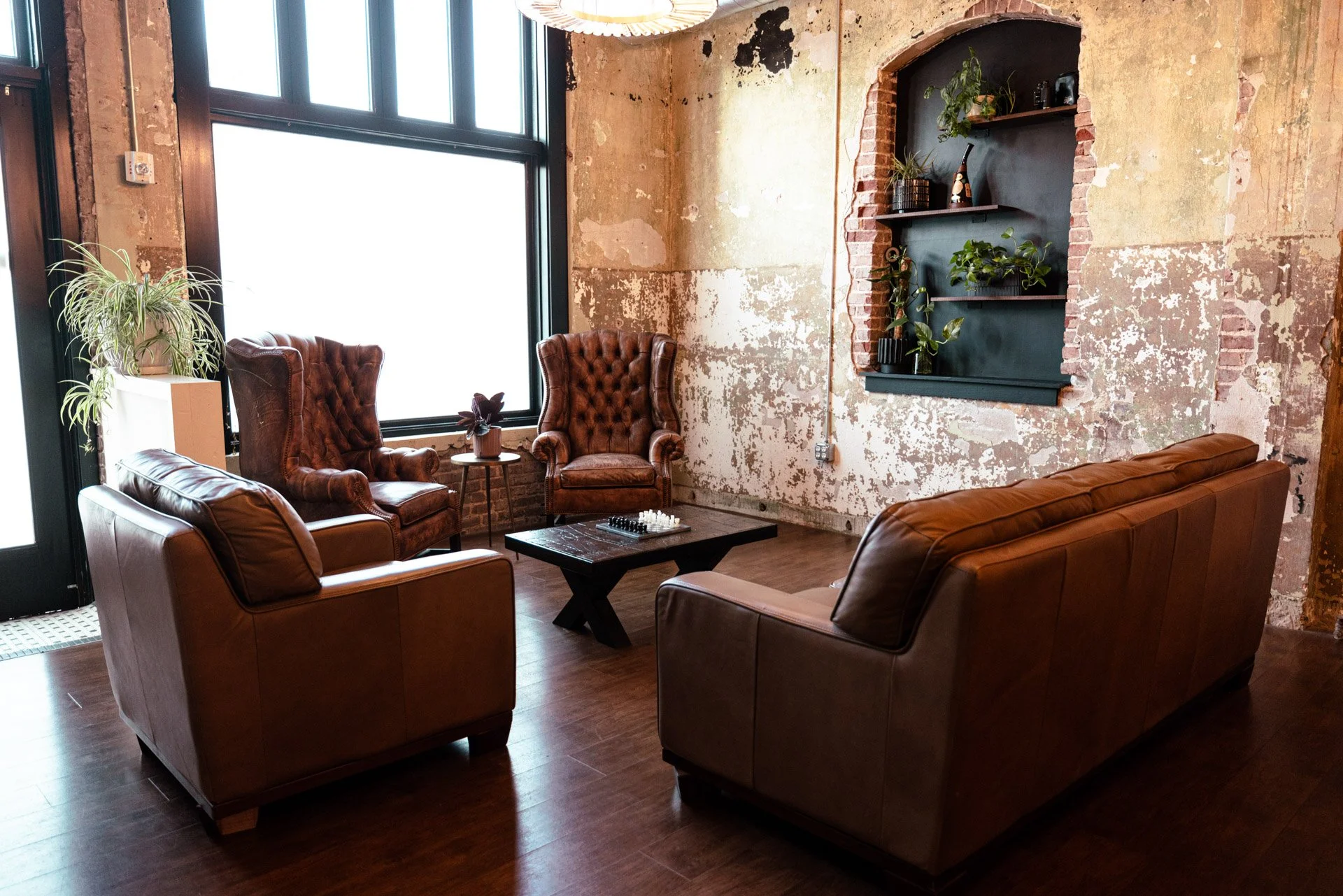 Interior of a rustic, renovated space with exposed brick and peeling paint walls, large windows, ceiling air ducts, and modern lighting fixtures, including a yellow leather sofa, a large flat-screen TV on a rolling stand, and a bar-height table with chairs.