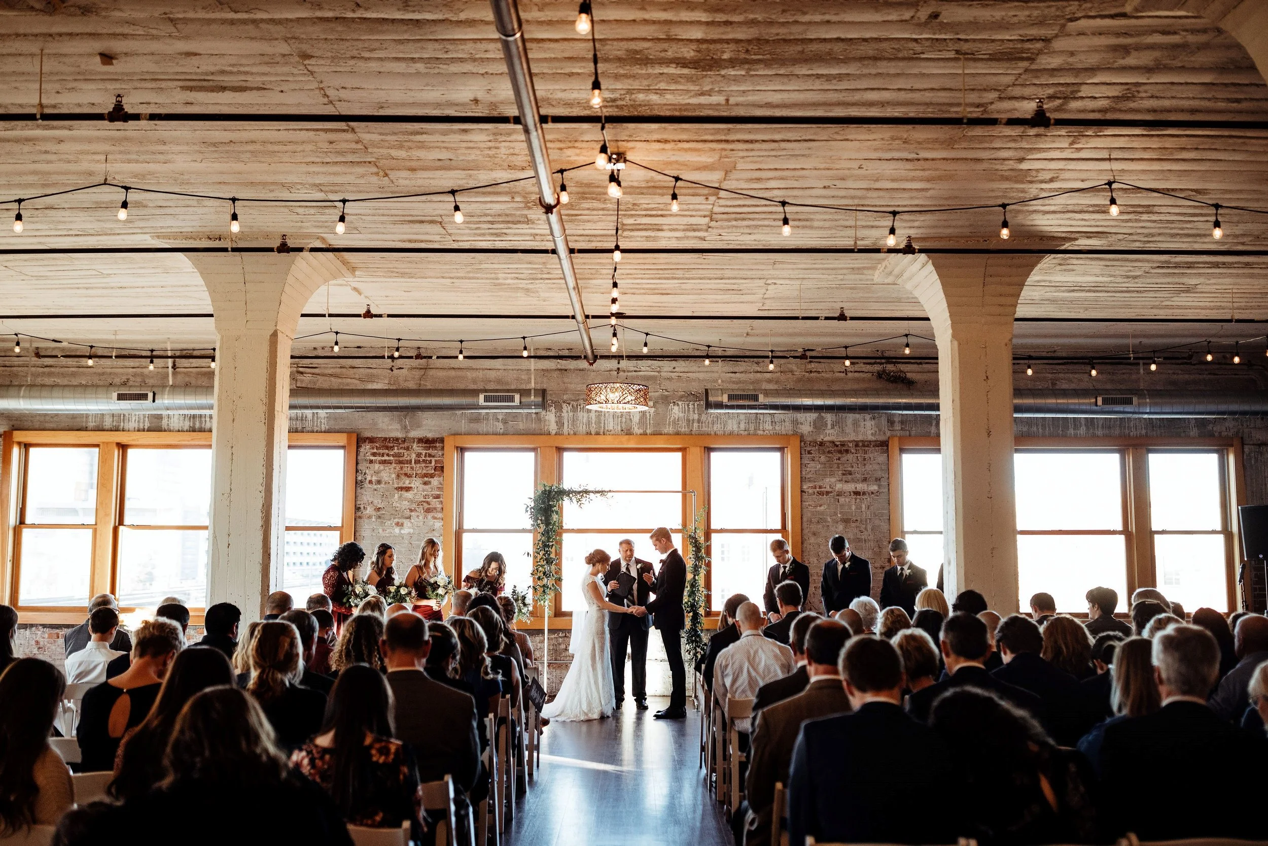 Indoor wedding ceremony with a bride and groom exchanging vows, officiant, bridesmaids, groomsmen, and seated guests in a rustic venue with exposed brick walls, large windows, and string lights.
