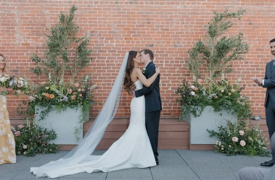 Bride and groom kiss during wedding ceremony outdoors, with two large floral arrangements and a brick wall in the background.